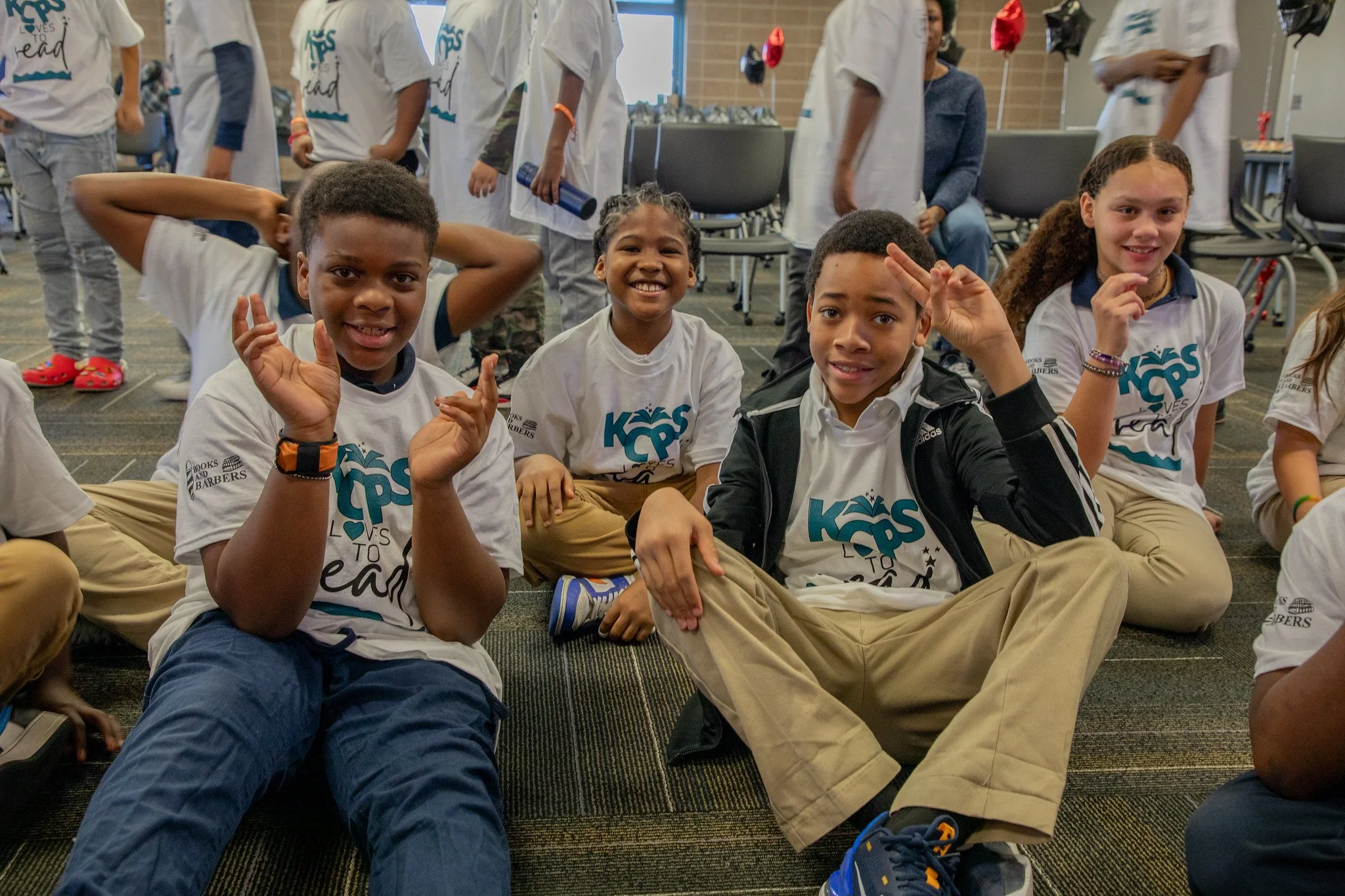 Smiling children sitting on the floor in a group at a school event, wearing matching T-shirts with a logo that says 'Kids Loves to Learn' and with balloons in the background.