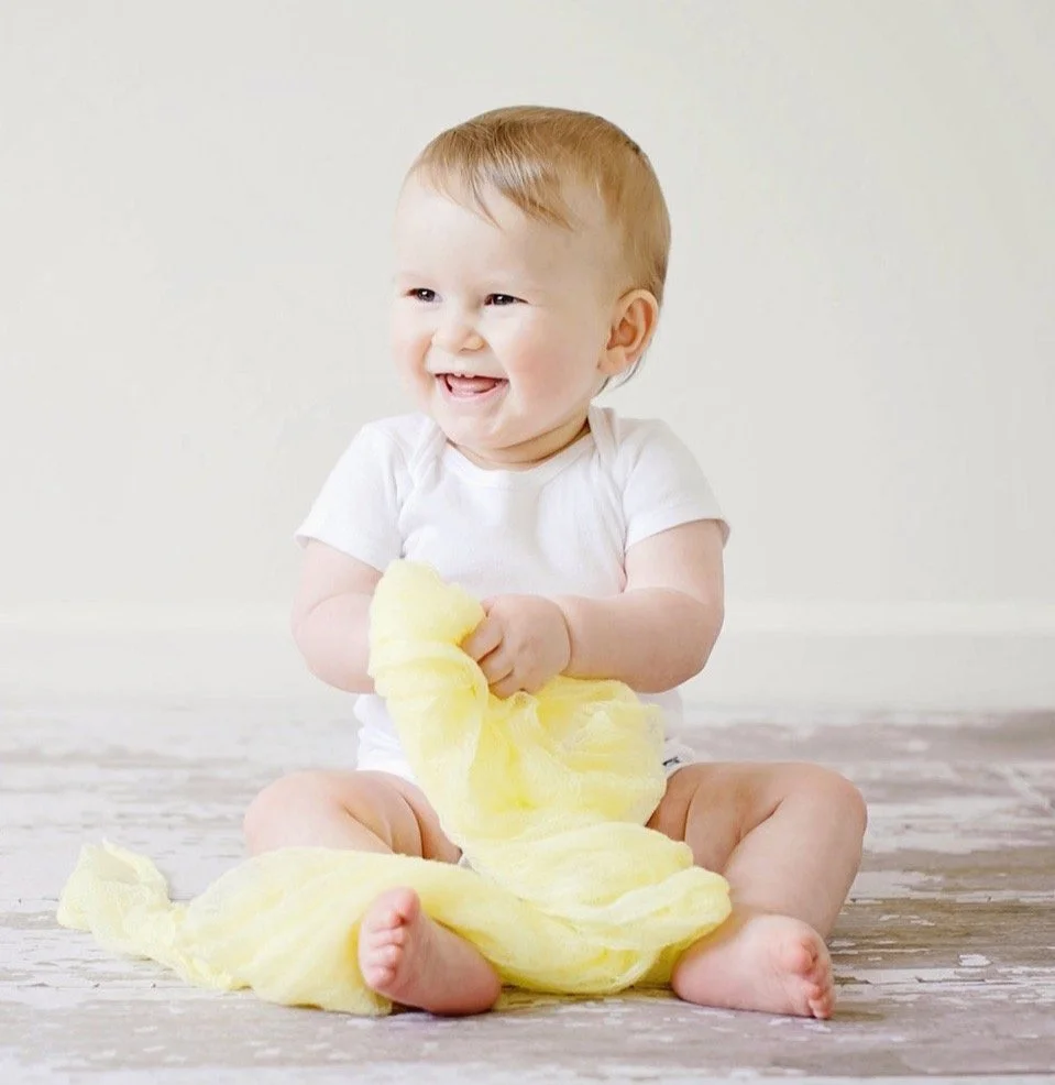 Smiling baby sitting on a wooden floor, holding yellow cloth, dressed in a white shirt.