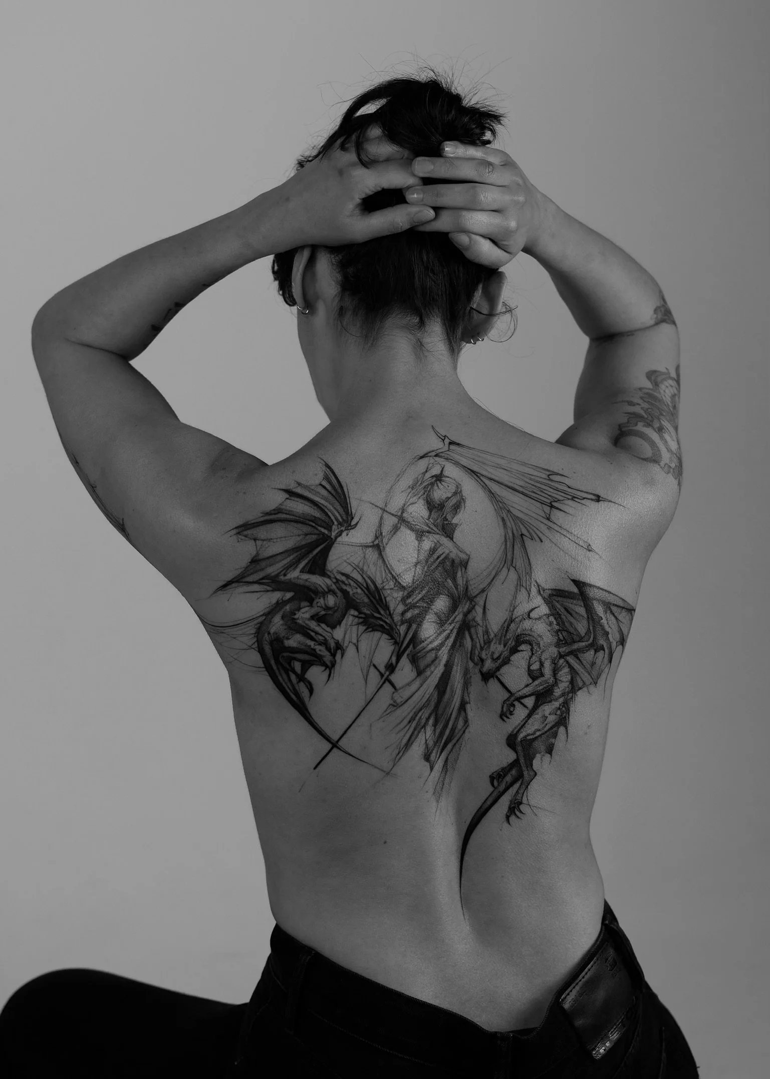 Back view of a person with short hair and multiple tattoos, sitting with one knee up. They are holding their head with hands, with elbows pointing outward.