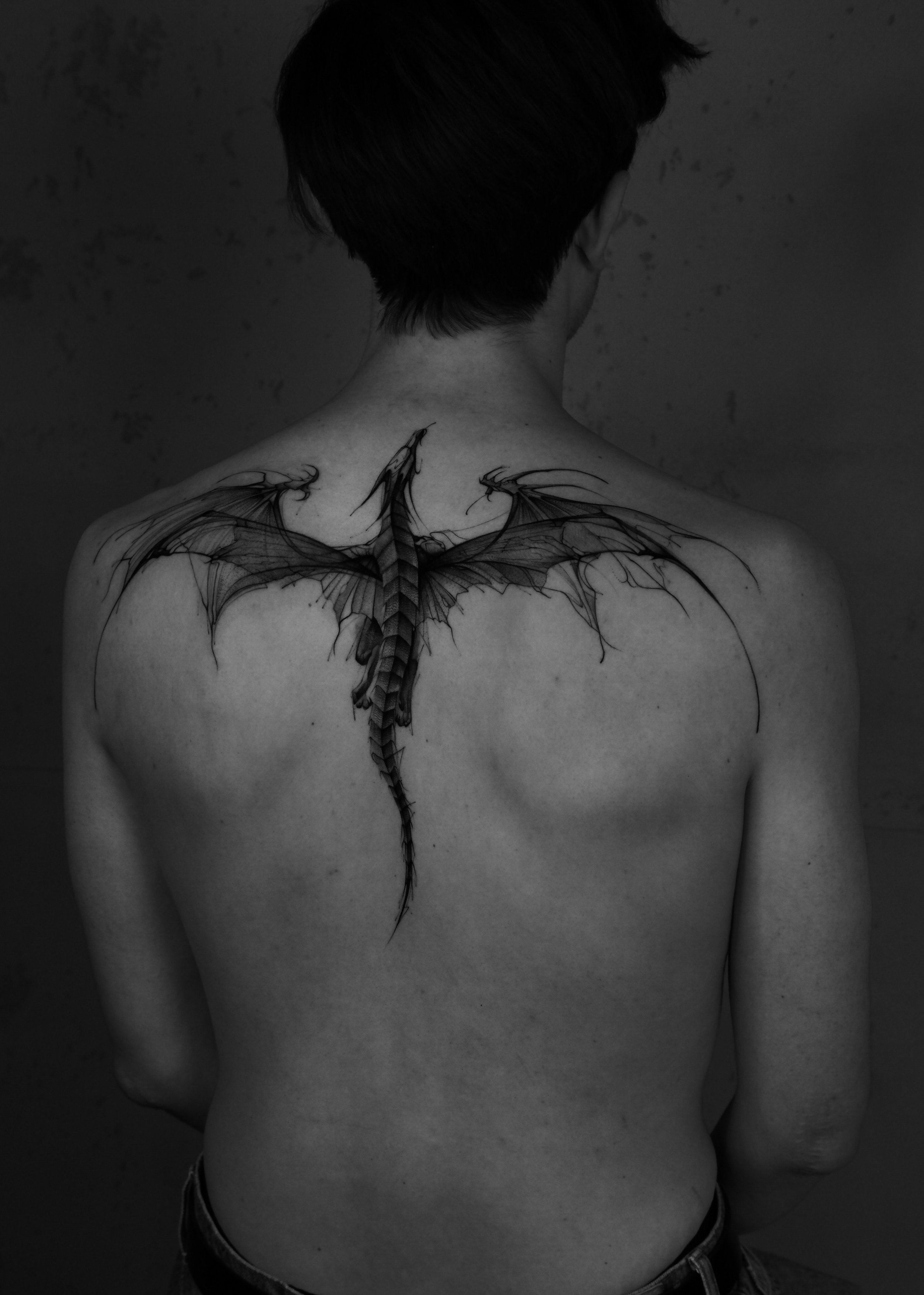 Back of a person with a dragon tattoo on their upper back and shoulders, showing only from the head to the hips, in black and white.