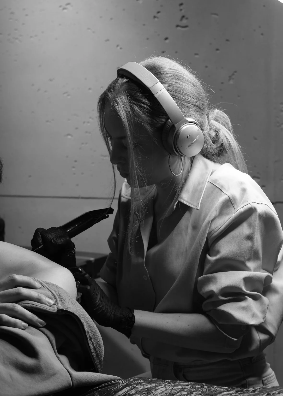 A woman in protective gloves and headphones is tattooing a person's arm with a tattoo machine.