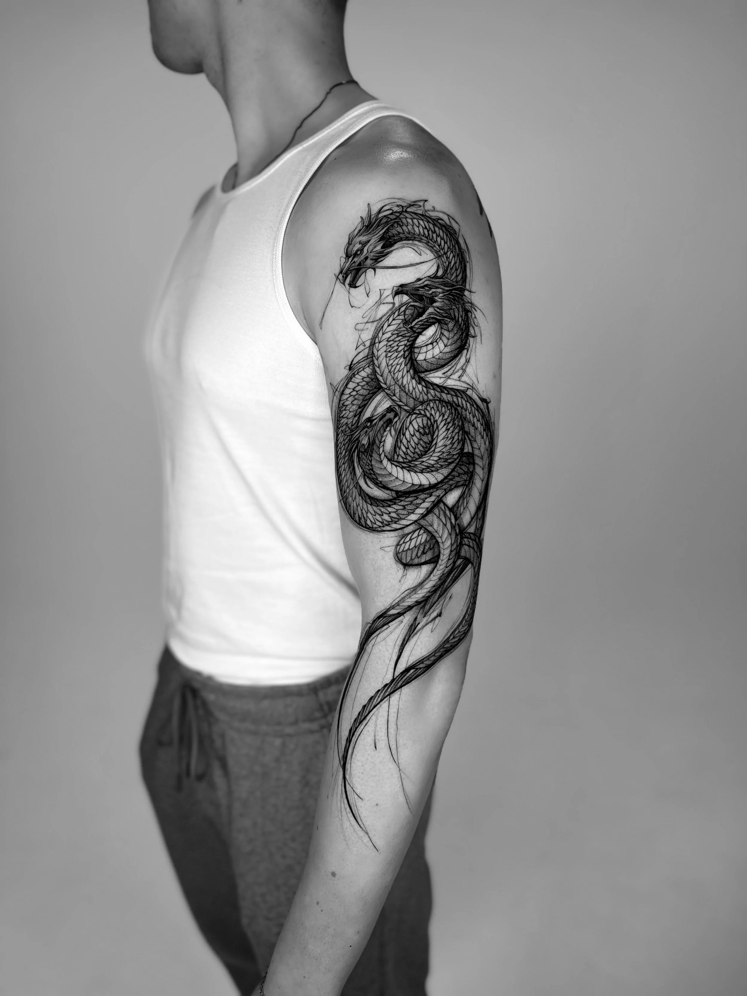 A person with a dragon tattoo on their upper arm, wearing a white sleeveless shirt and gray shorts, standing against a plain background.