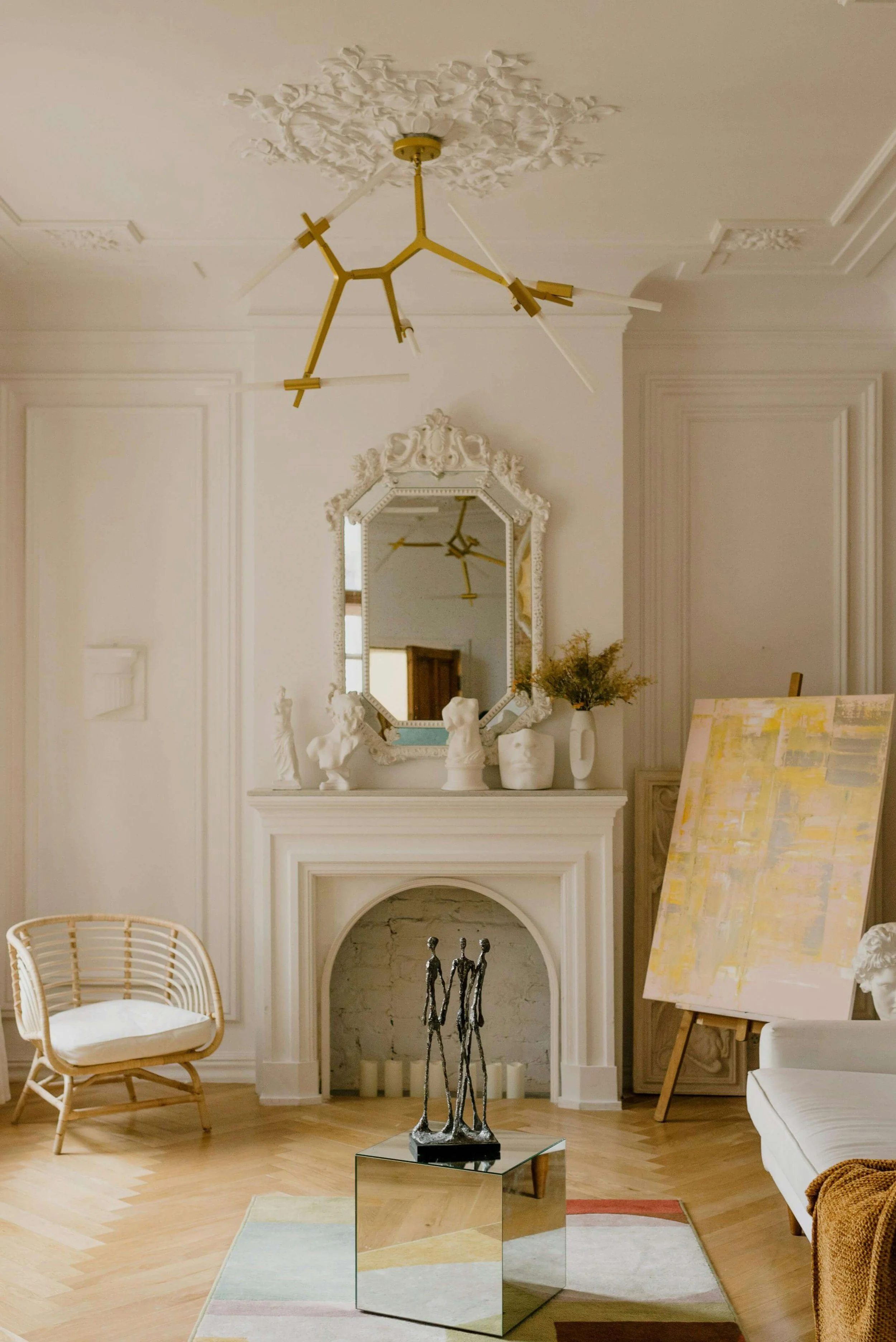 cream colored living room with geometric gold ceiling lught, fireplace, painting on easel in the corner and mirrow over the fireplace