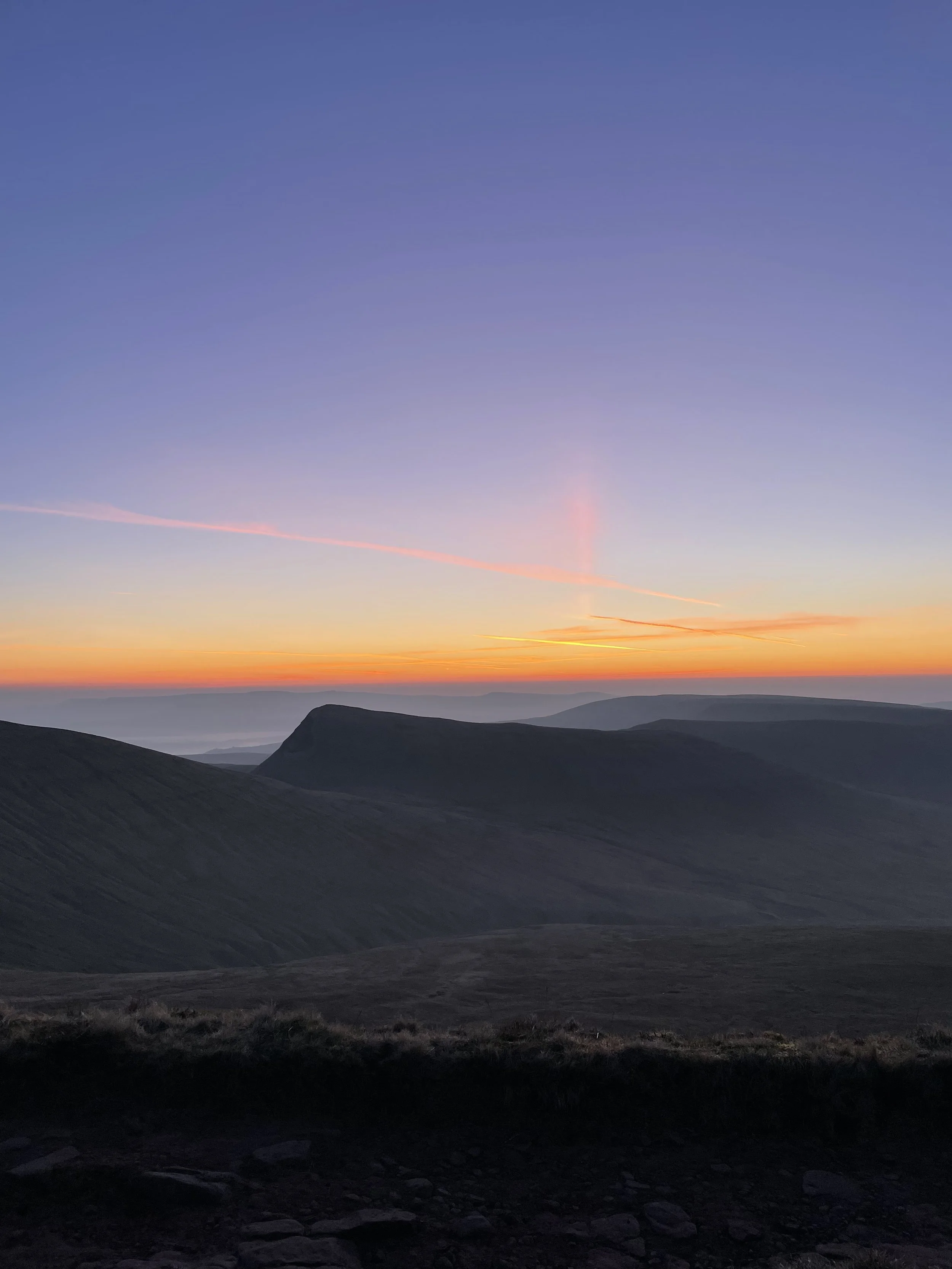 A scenic view of distant rolling hills or mountains at sunset with a sky painted in shades of purple, pink, orange, and yellow.