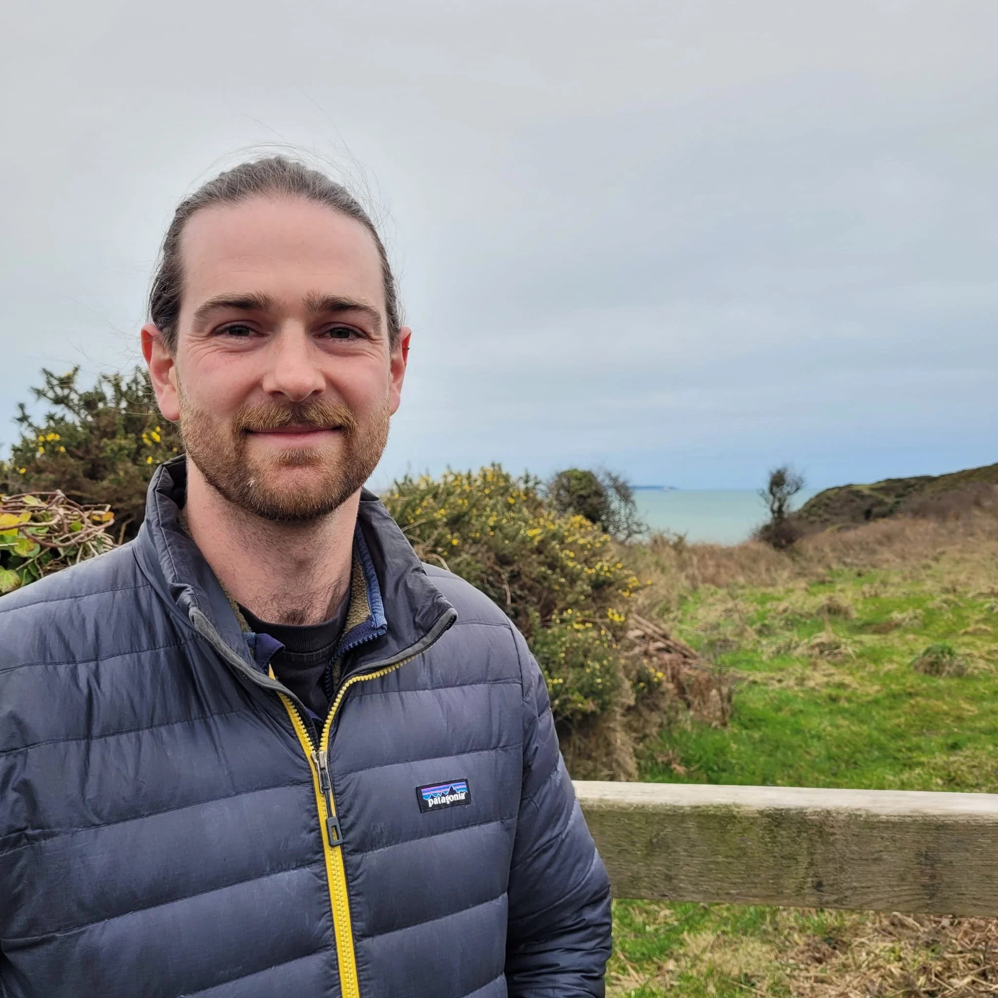 Close-up of a smiling man with a beard and long hair tied back, wearing a dark blue Patagonia puffer jacket, outdoors near a body of water and green trees.