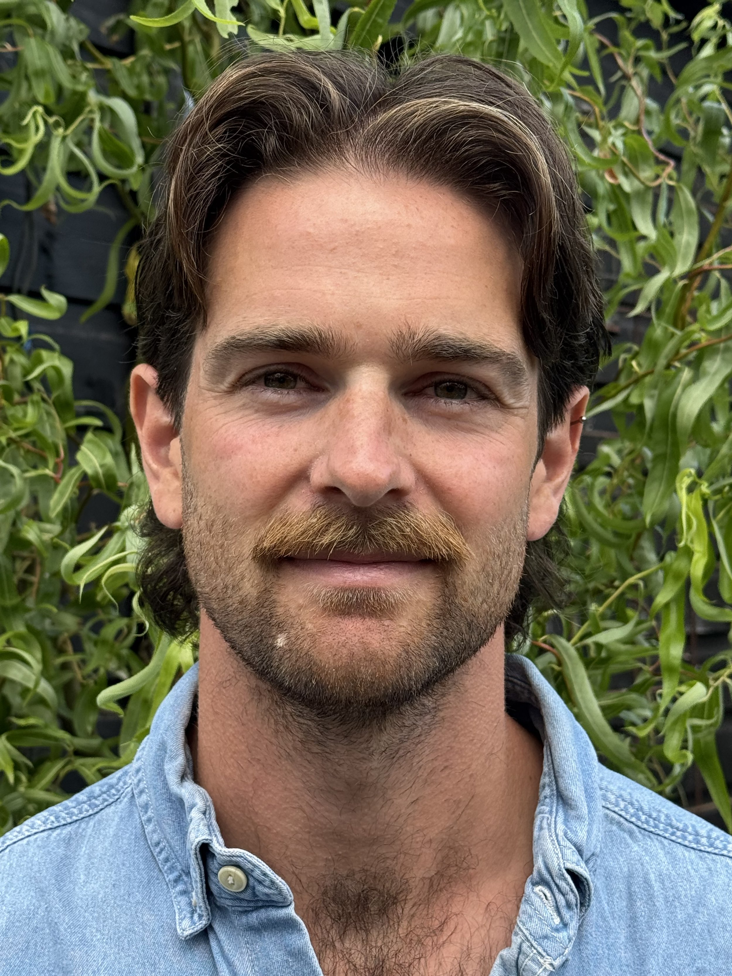 Close-up of a man with waving brown hair and a beard, wearing a light blue denim shirt, standing in front of green leafy plants.