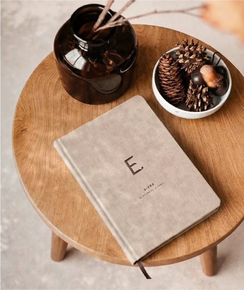Un livre beige avec la lettre 'E' sur la couverture, placé sur une table en bois ronde. À côté, un pot en verre sombre avec des bâtons d'encens et un bol blanc contenant des pommes de pin, des châtaignes et d'autres fruits secs.