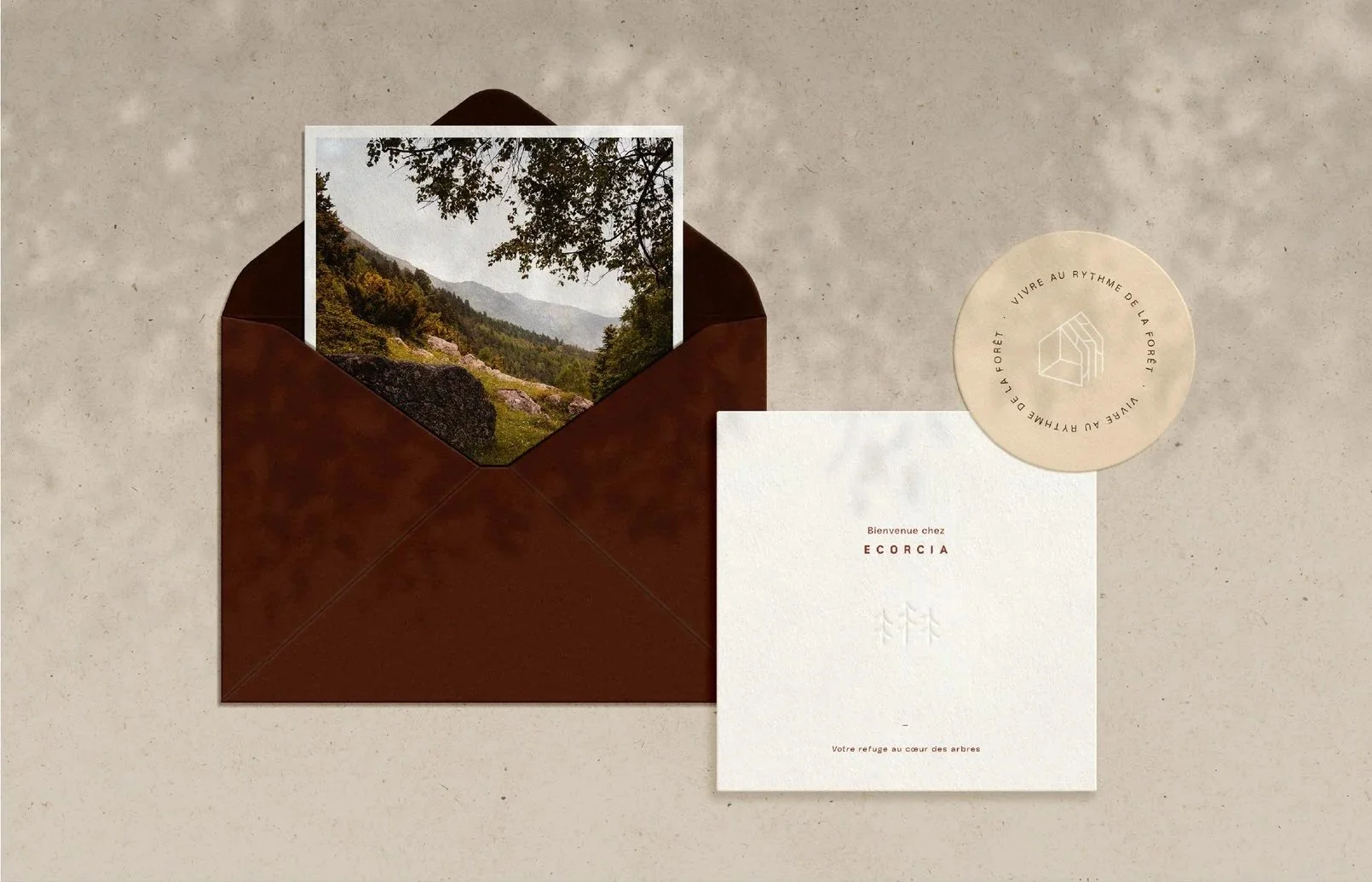 Une enveloppe marron contenant une photo de paysage montagneux avec des arbres et des collines vertes, accompagnée d'une carte blanche avec un texte en français et un cylindre en papier beige avec logo et inscriptions.