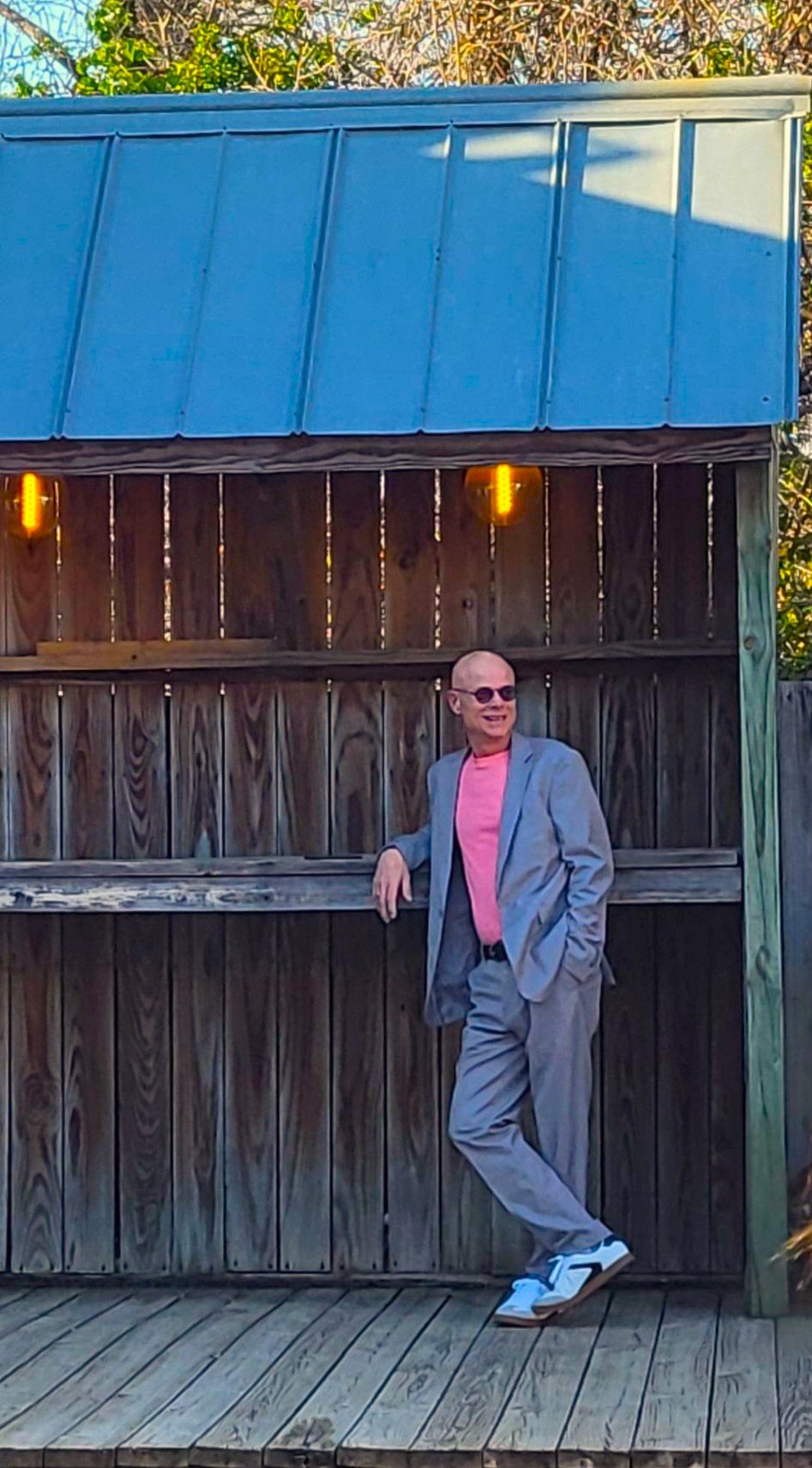 A man in a light gray suit with a pink shirt and sunglasses, standing with one hand on a wooden bar or counter, smiling, outdoors on a wooden deck near a rustic wooden fence with string lights.