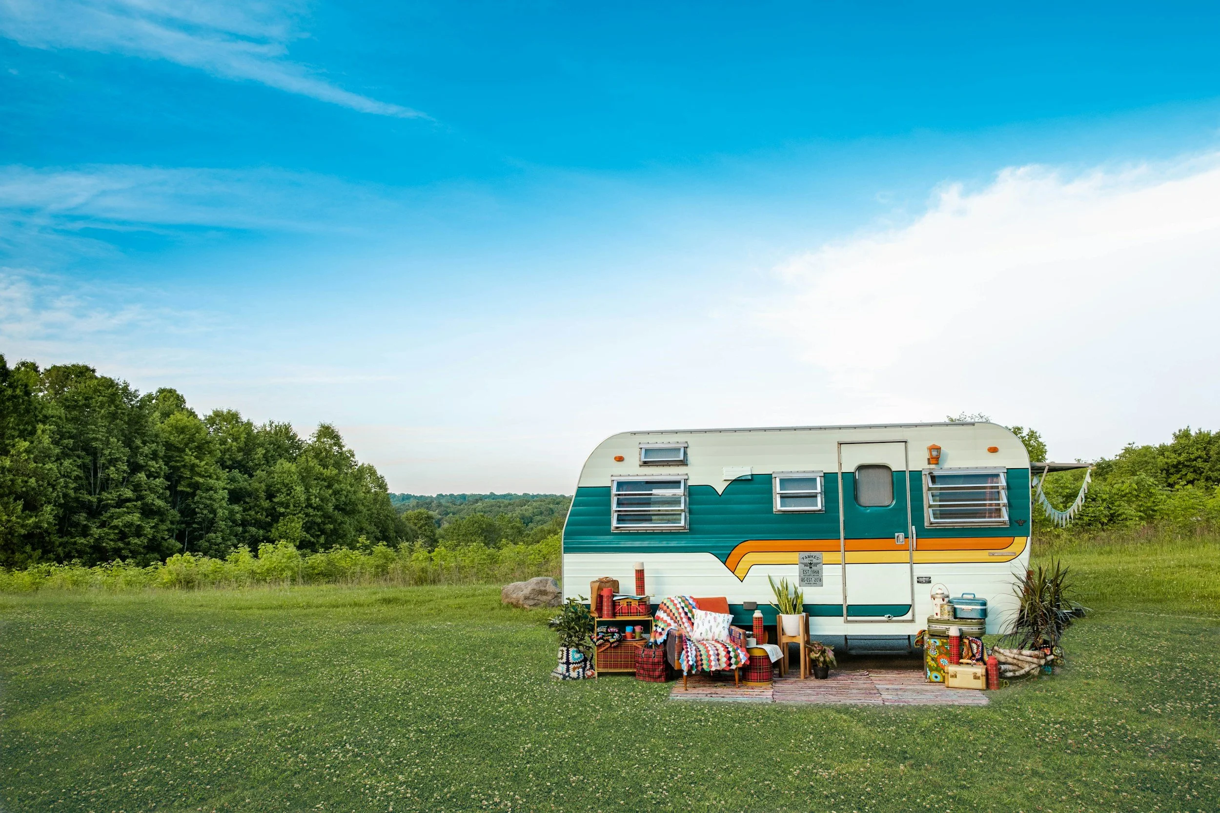 A vintage camper trailer parked in a grassy field with outdoor furniture and decorative items, surrounded by trees under a blue sky.