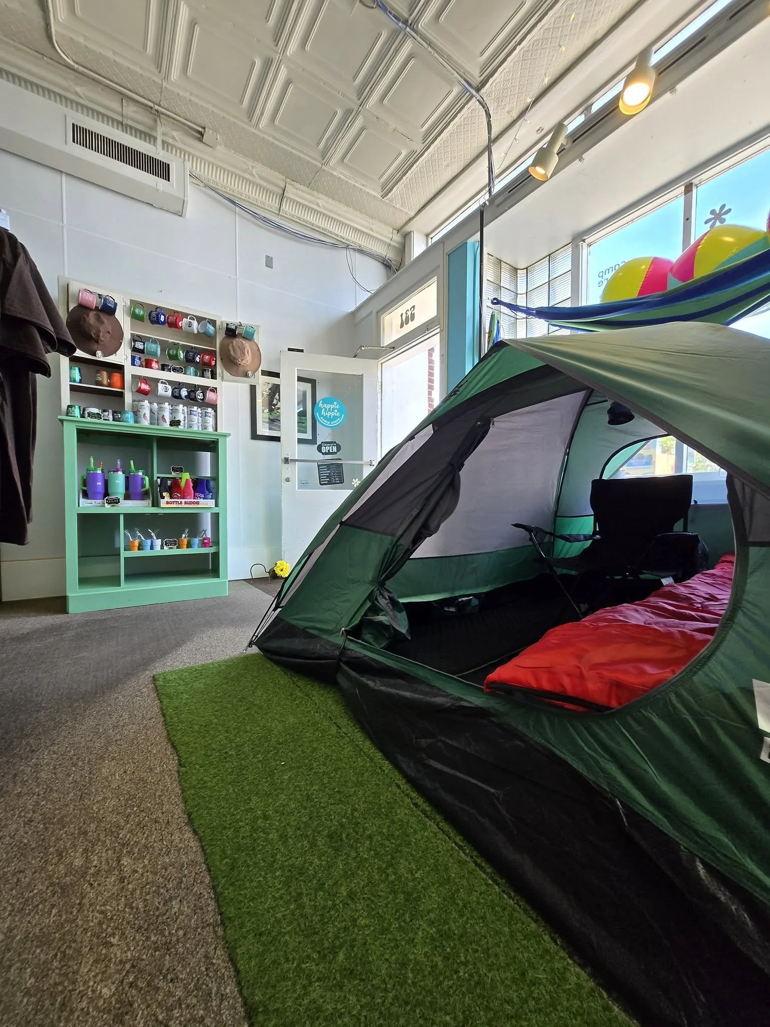 Indoor space with a green camping tent with red bedding, a small green shelf holding colorful bottles, a wall with mug and hat display, large windows with colorful balls, and a small patch of green artificial grass.