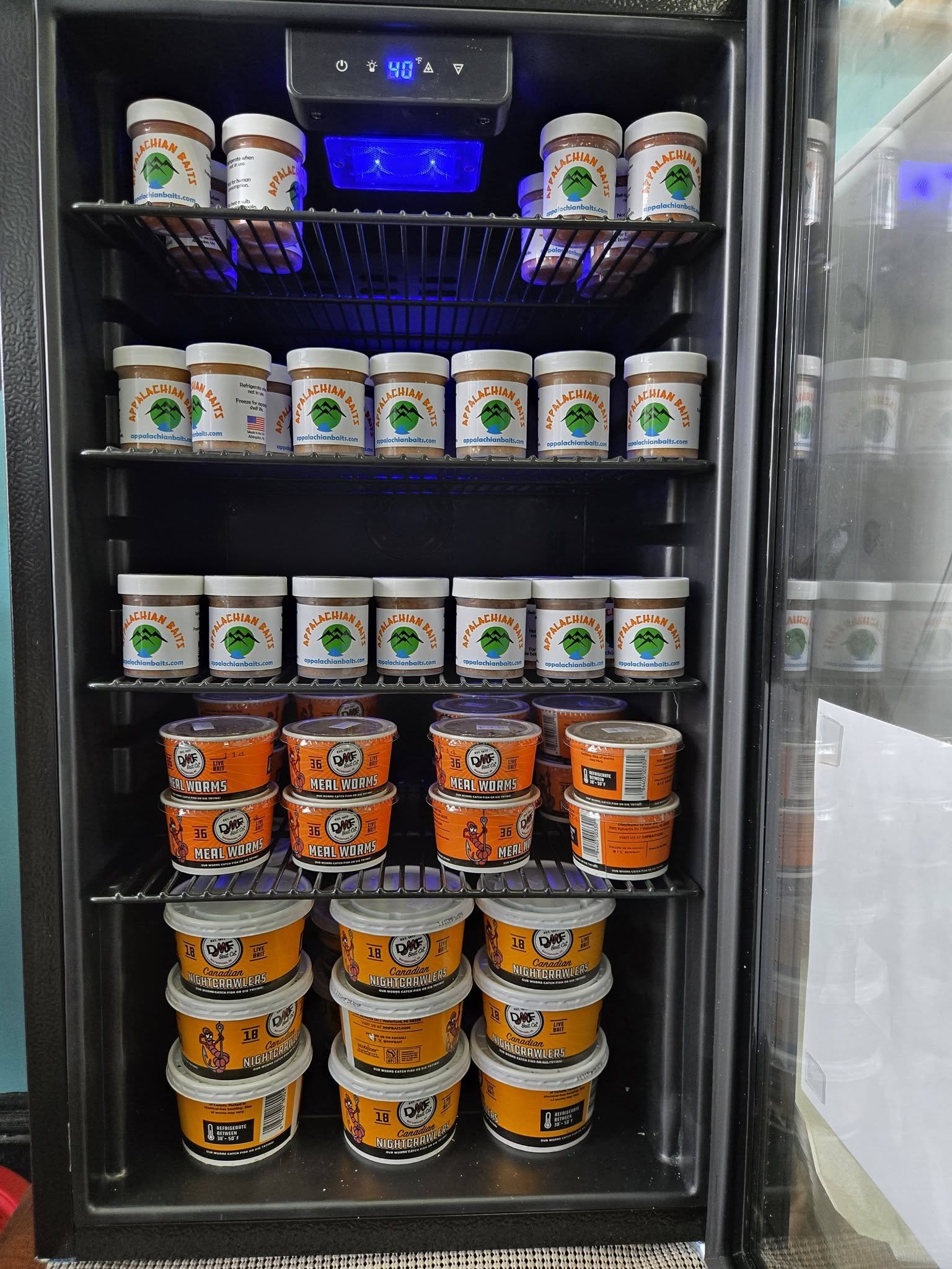 A refrigerator containing multiple containers of bait, specifically Canadian Nightcrawlers, with orange labels, black and white lids, and a logo with a cartoon fish. The refrigerator has a digital temperature display showing 40°F, and blue LED lighti