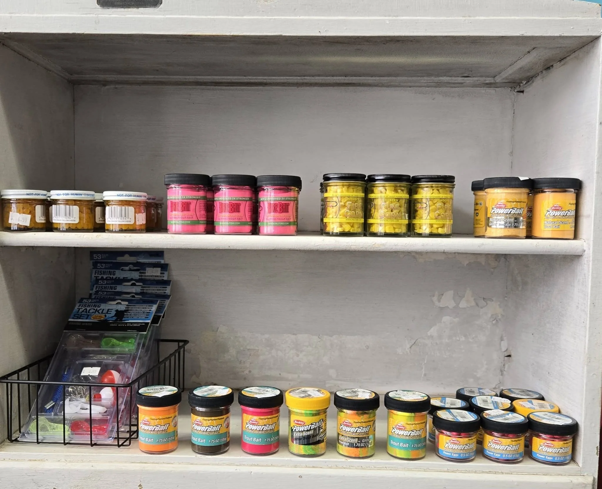 Store shelf with fishing bait containers, some jars and tins, and packaged fishing tackle below.