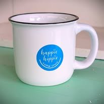 White enamel mug with blue logo that says 'happie frewpie' on a light green surface.