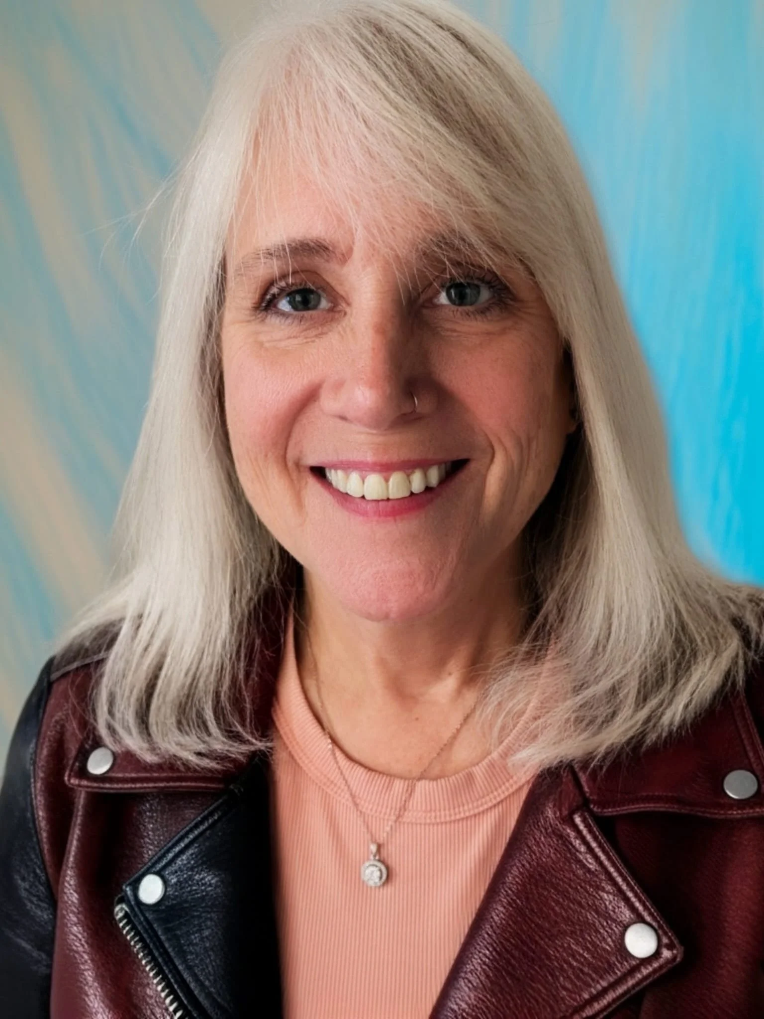 Close-up of a smiling woman with platinum blonde hair, wearing a pink ribbed top, a maroon leather jacket, and a silver necklace, against a blue background.