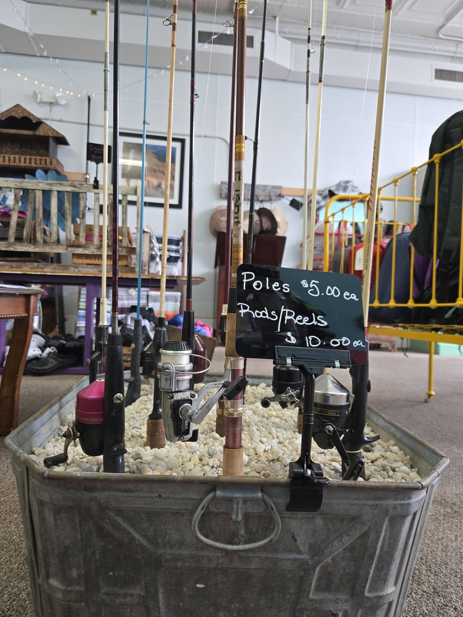 Gently used fishing poles in a metal cart with rocks, a sign indicating prices, and a wooden shelf in the background.