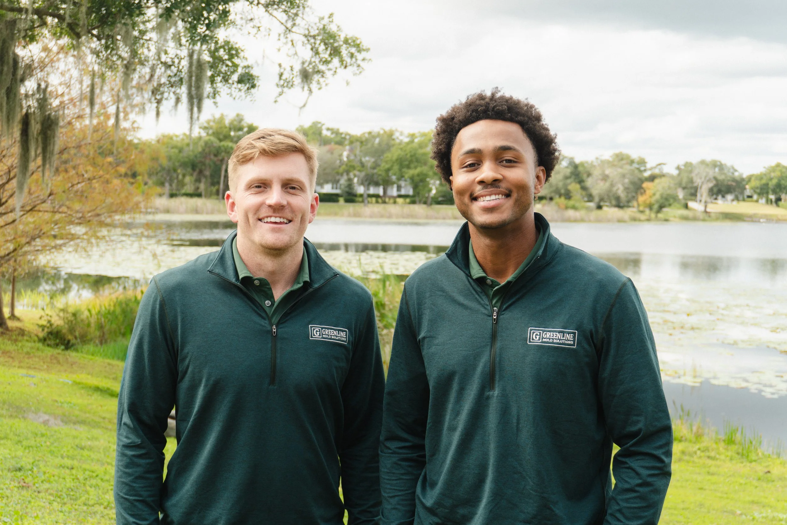 Two smiling men standing outdoors near a lake with trees in the background, wearing matching dark green jackets with a logo that says 'Greenline Mold Solutions'.