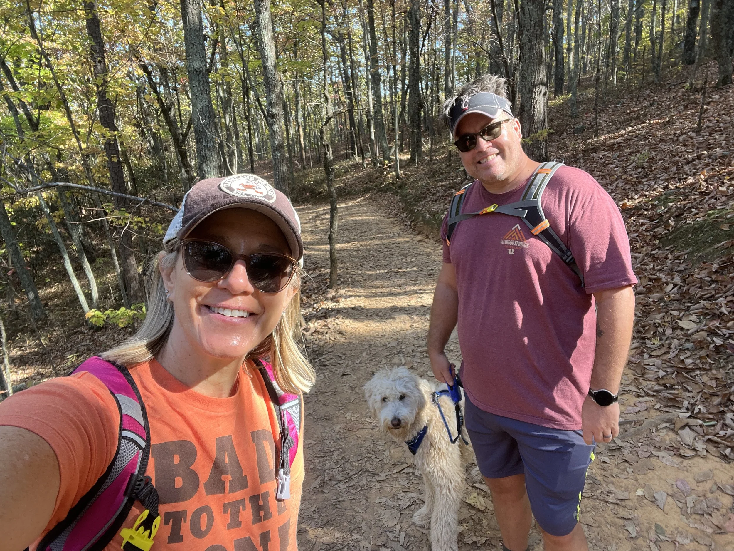 A woman and a man with sunglasses taking a selfie during a hike in a wooded area, with a small white dog on a leash between them.