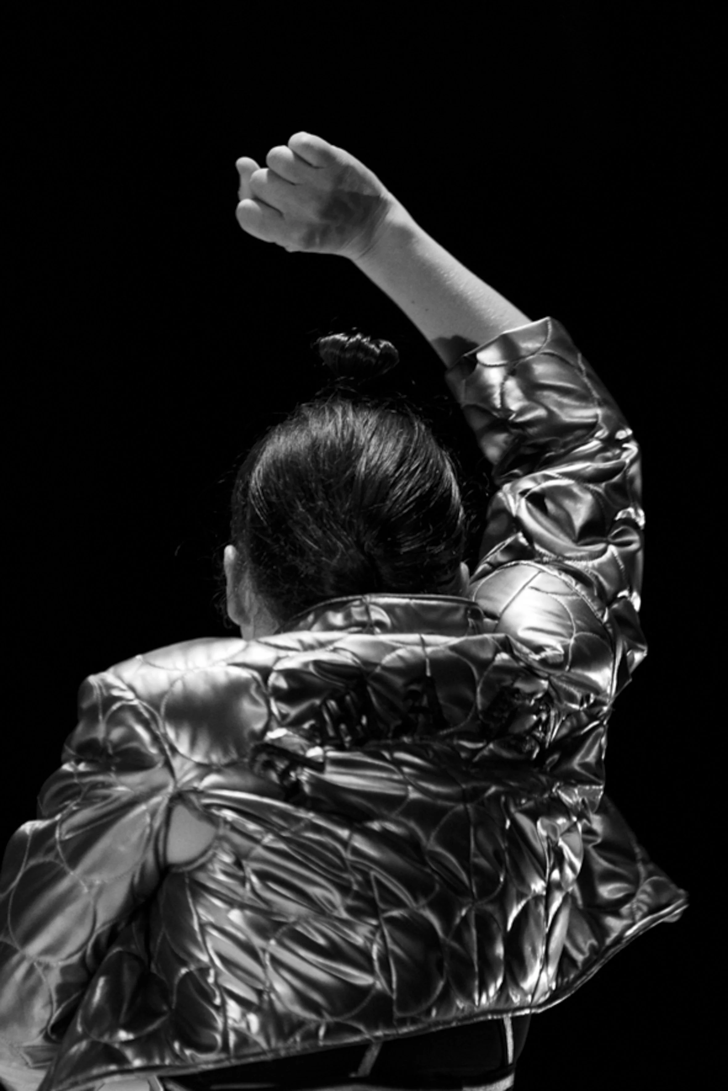 Une personne vue de dos, portant une veste en cuir brillant, soulève un bras en l'air dans un espace sombre.