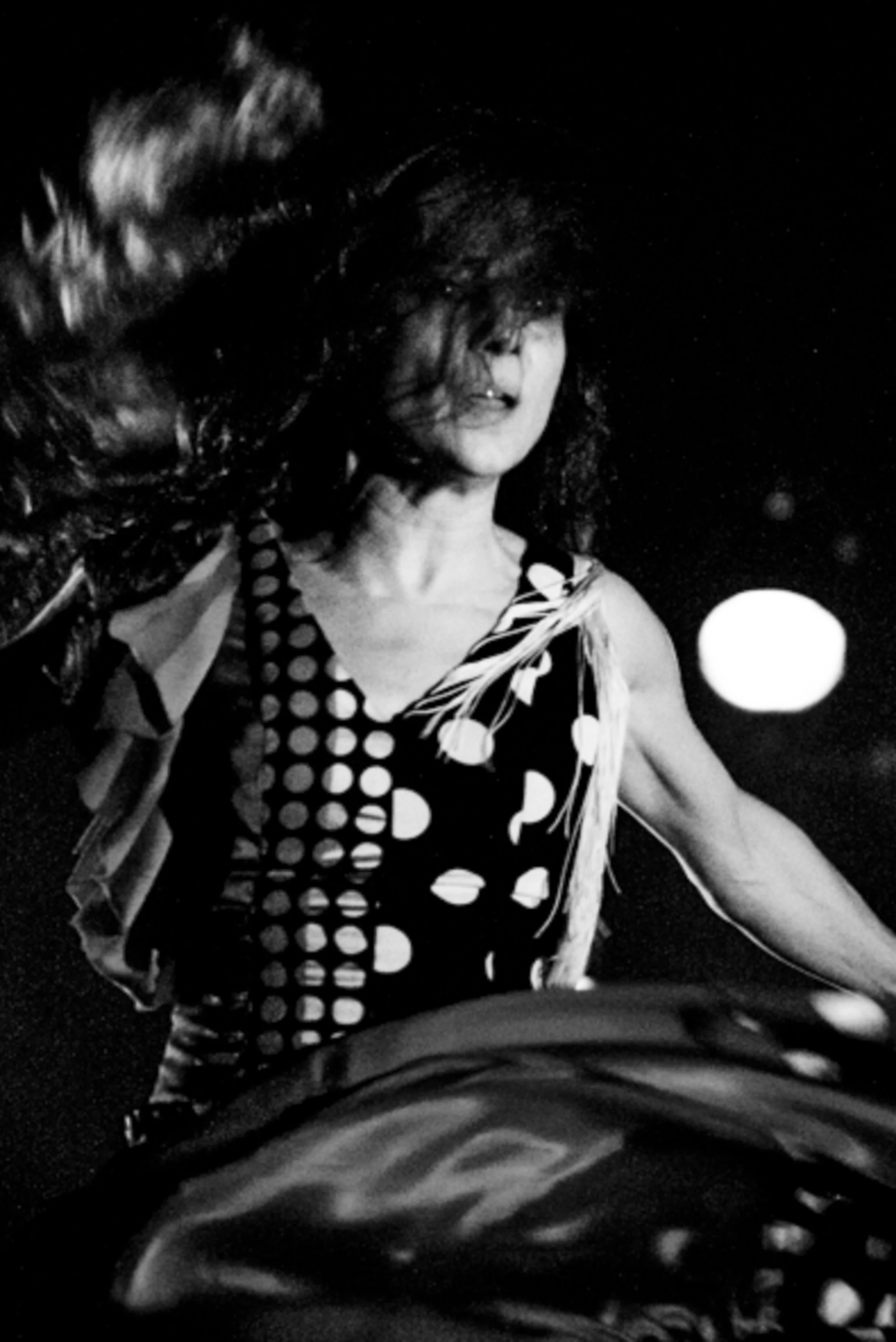 Une femme en noir et blanc avec des cheveux en mouvement, portant une robe à motifs géométriques, dans un décor sombre avec une lumière ronde en arrière-plan.
