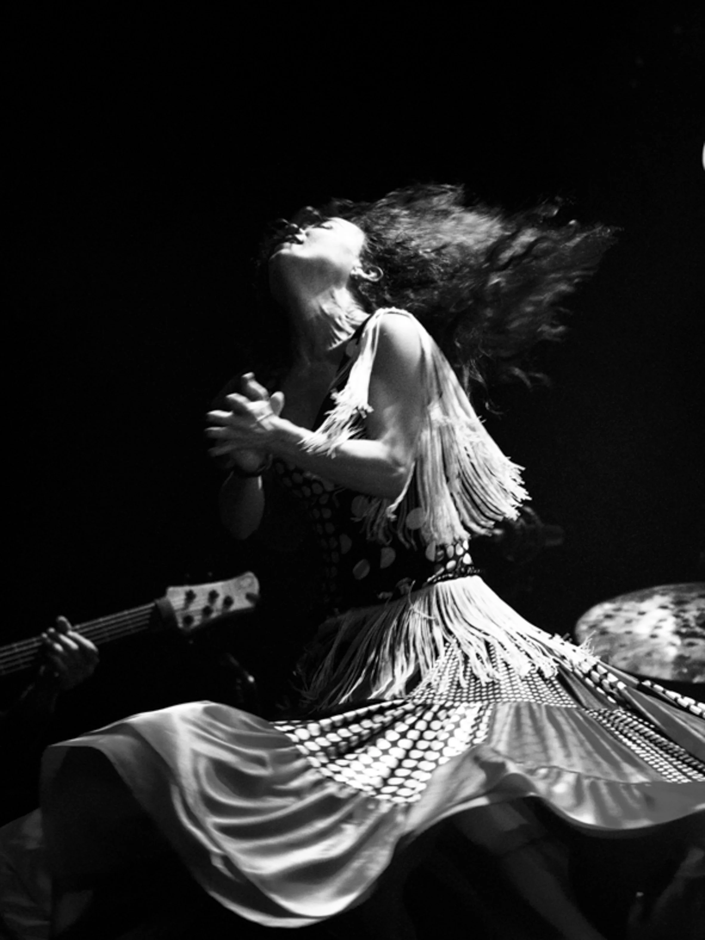 Une femme en robe fluide expressive, les yeux fermés, tourne la tête en arrière avec ses cheveux en mouvement, sur fond noir.