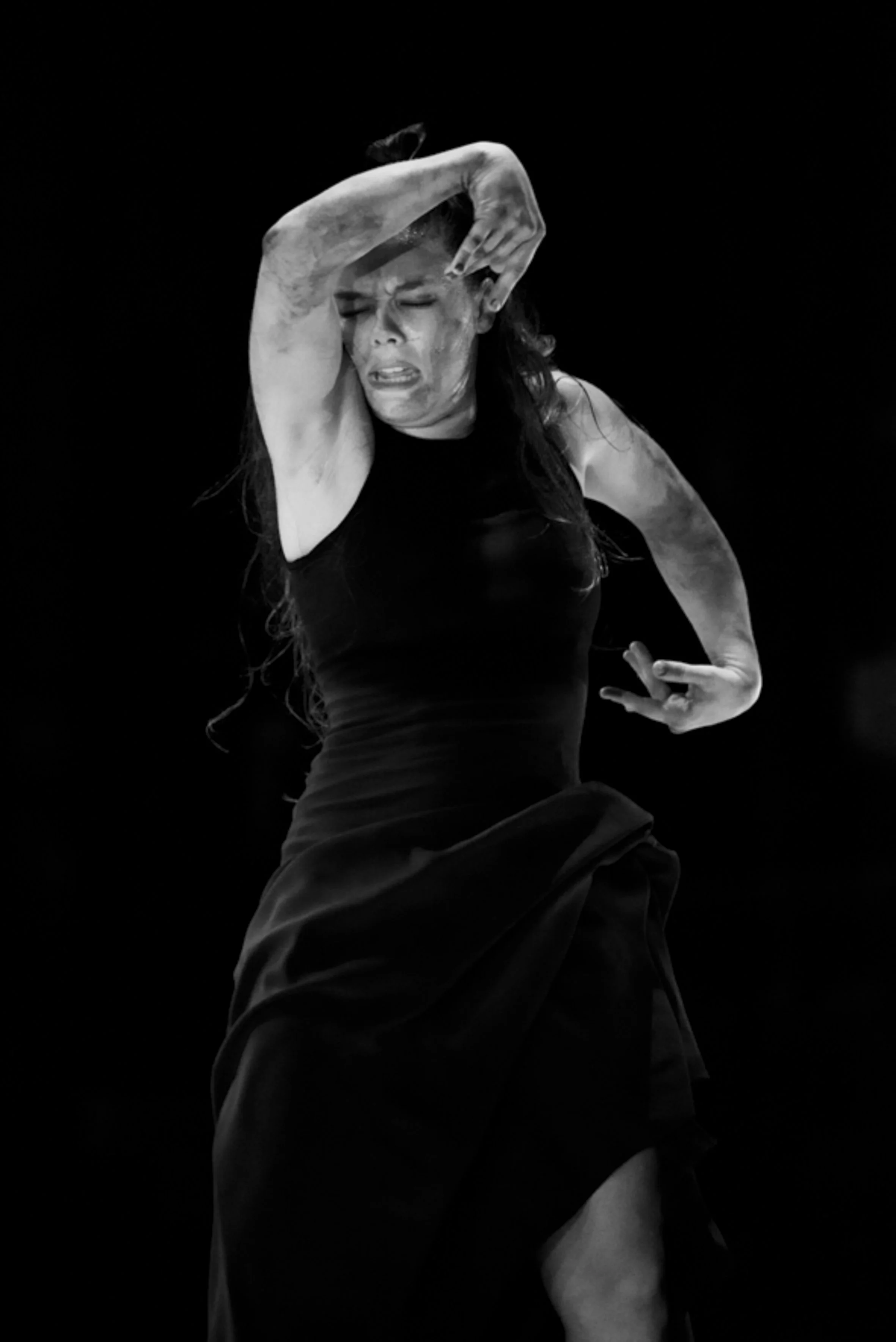 Une femme en noir tient une posture expressive, avec une expression intense sur son visage, contre un fond noir.