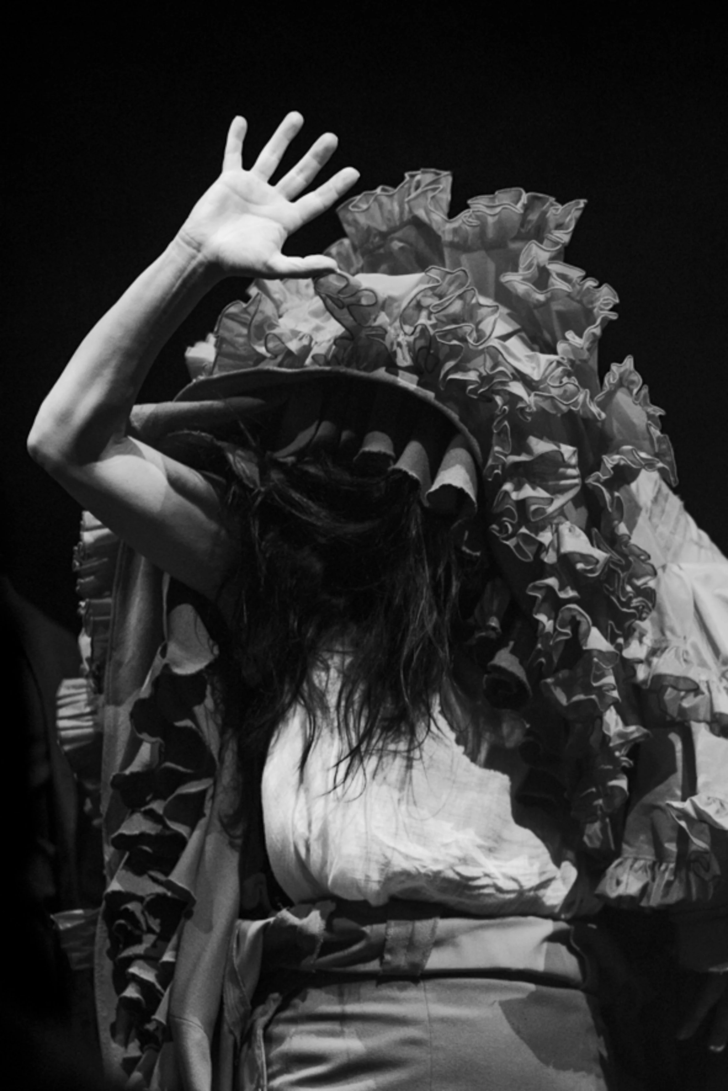 Une personne portant un costume extravagant avec un grand chapeau décoré de volants, couvrant partiellement son visage, avec une main levée en salut.