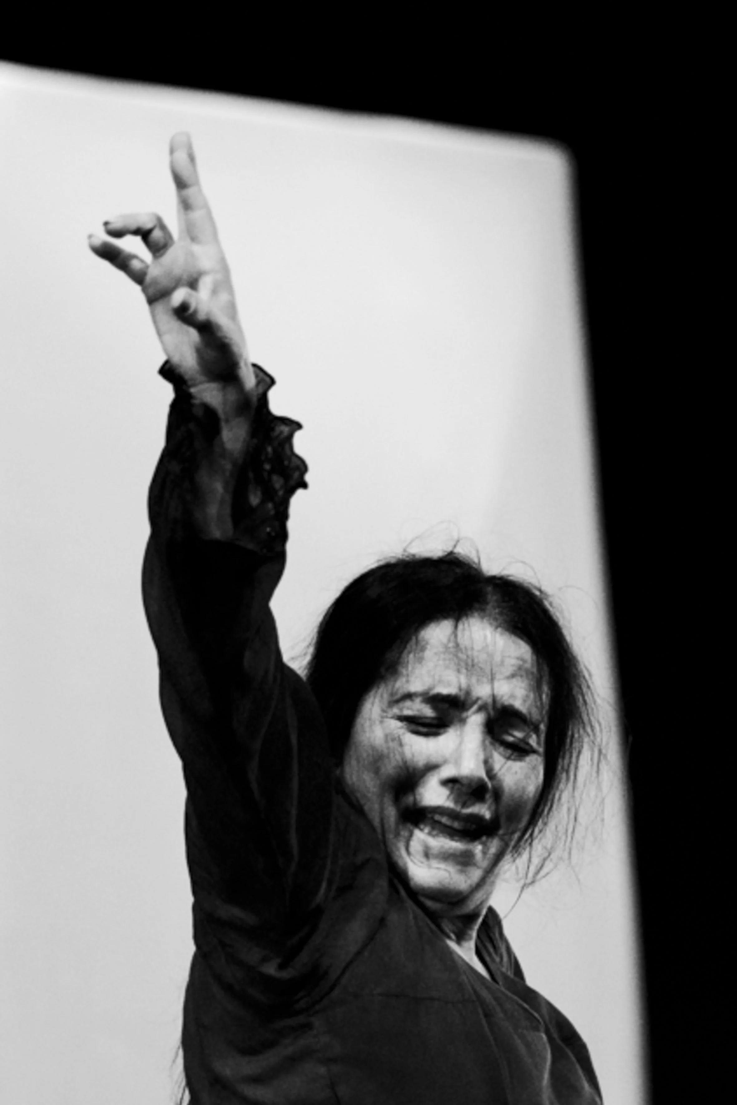 Une femme souriante, avec des cheveux noirs, la tête inclinée, semble faire une performance artistique ou dans une scène dramatique, en noir et blanc.