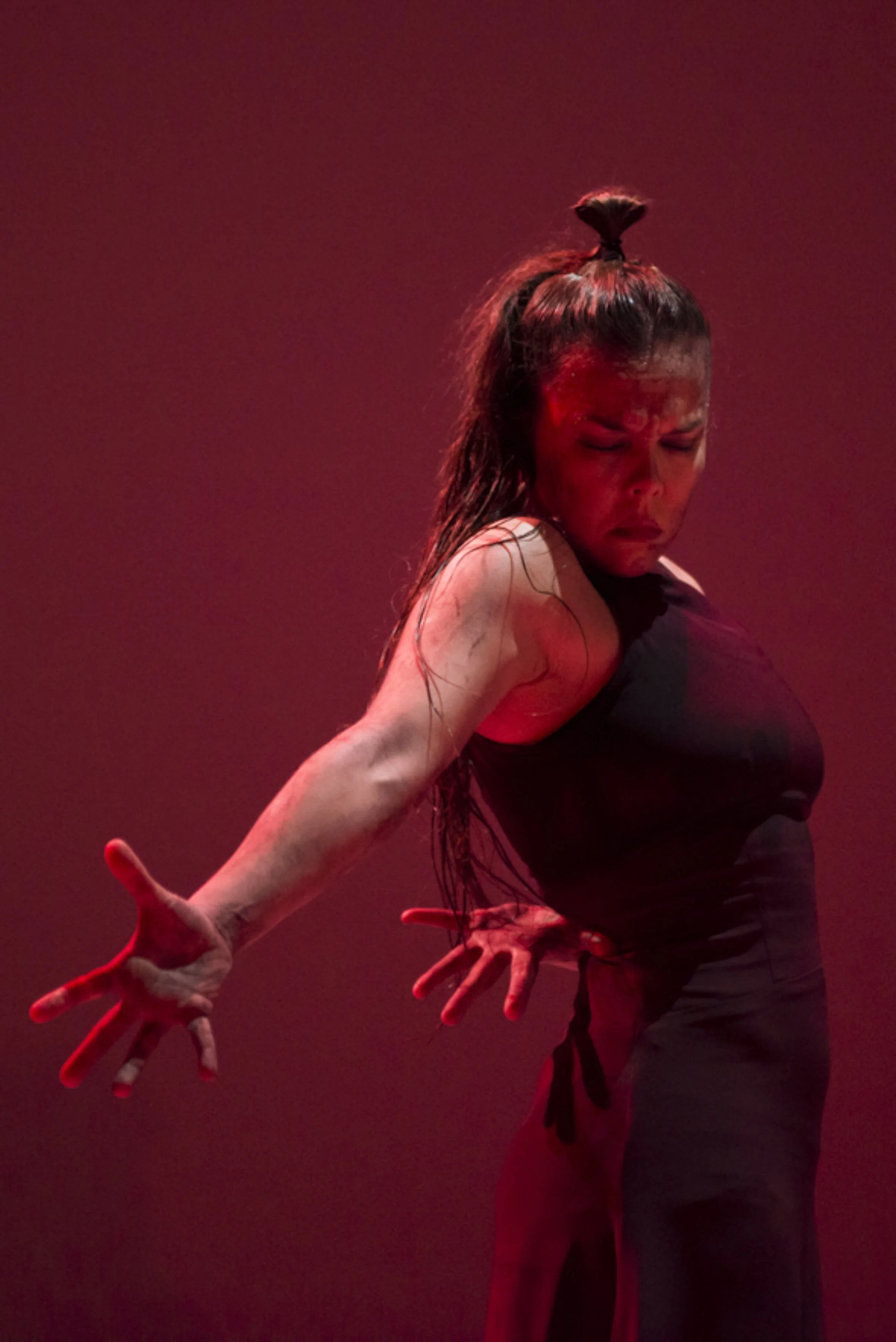 Une femme danse dans une pose expressive avec un fond rouge, portant une veste noire et une jupe longue, cheveux mouillés, expression intense.