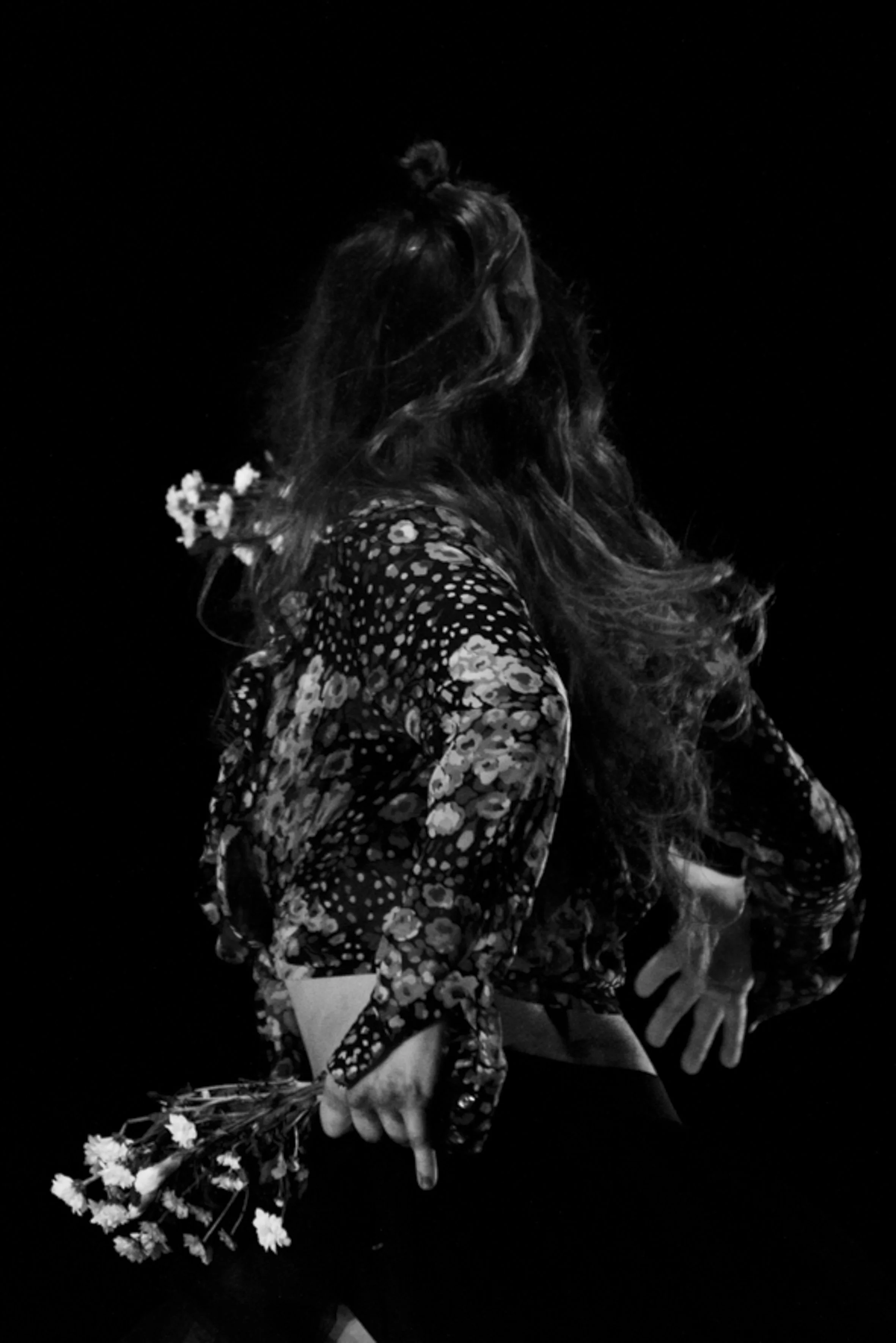 Femme avec cheveux longs et bouclés portant un vêtement à motifs floraux, tenant un petit bouquet de fleurs, dans un décor sombre.