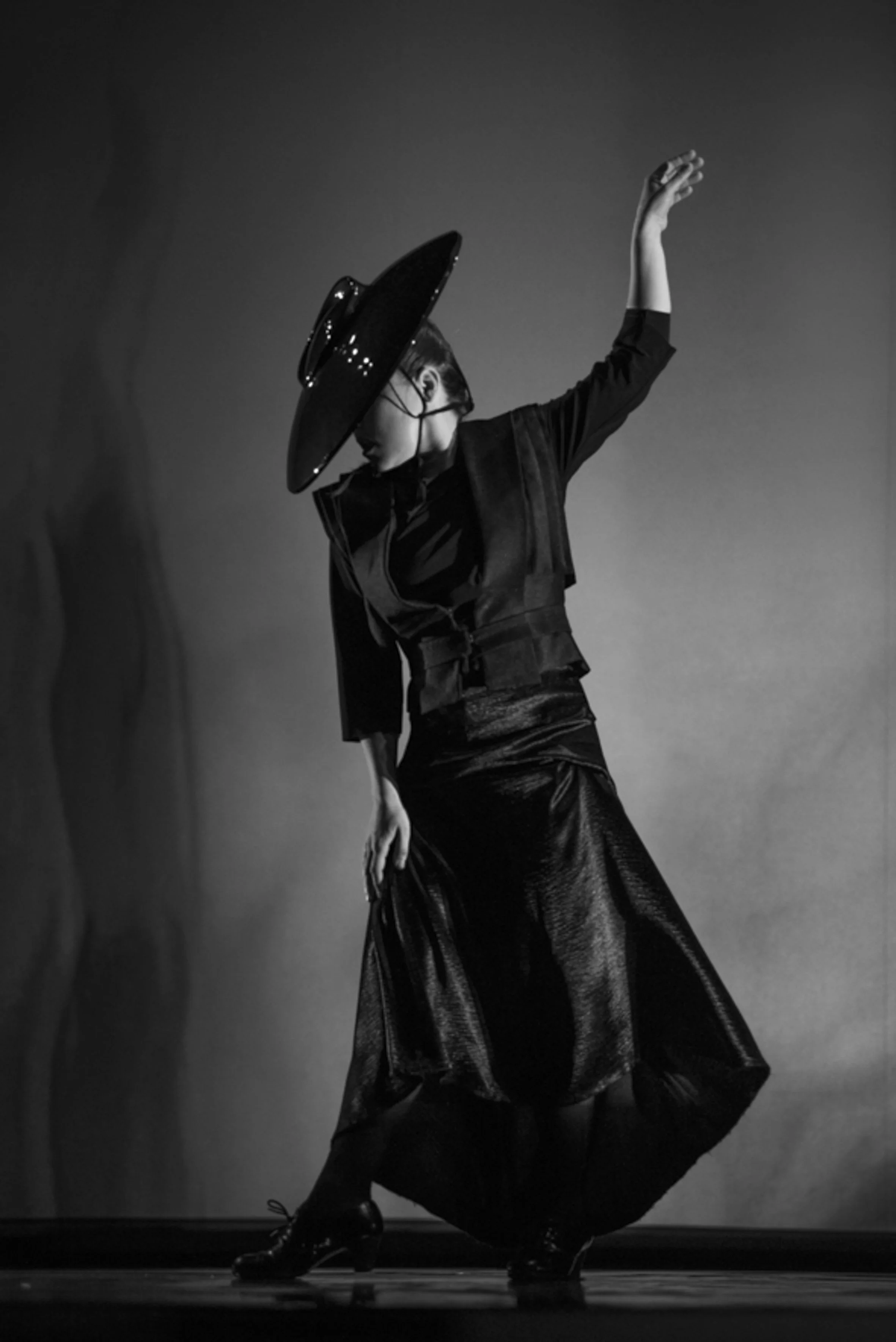Une femme portant un chapeau noir large, une veste sombre et une longue jupe en cuir noire, en train de danser sur scène, vue en noir et blanc.