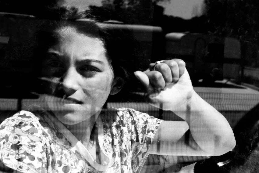 Une jeune femme regarde à travers une vitre, en noir et blanc, avec une expression sérieuse, portant un haut à motifs floraux.