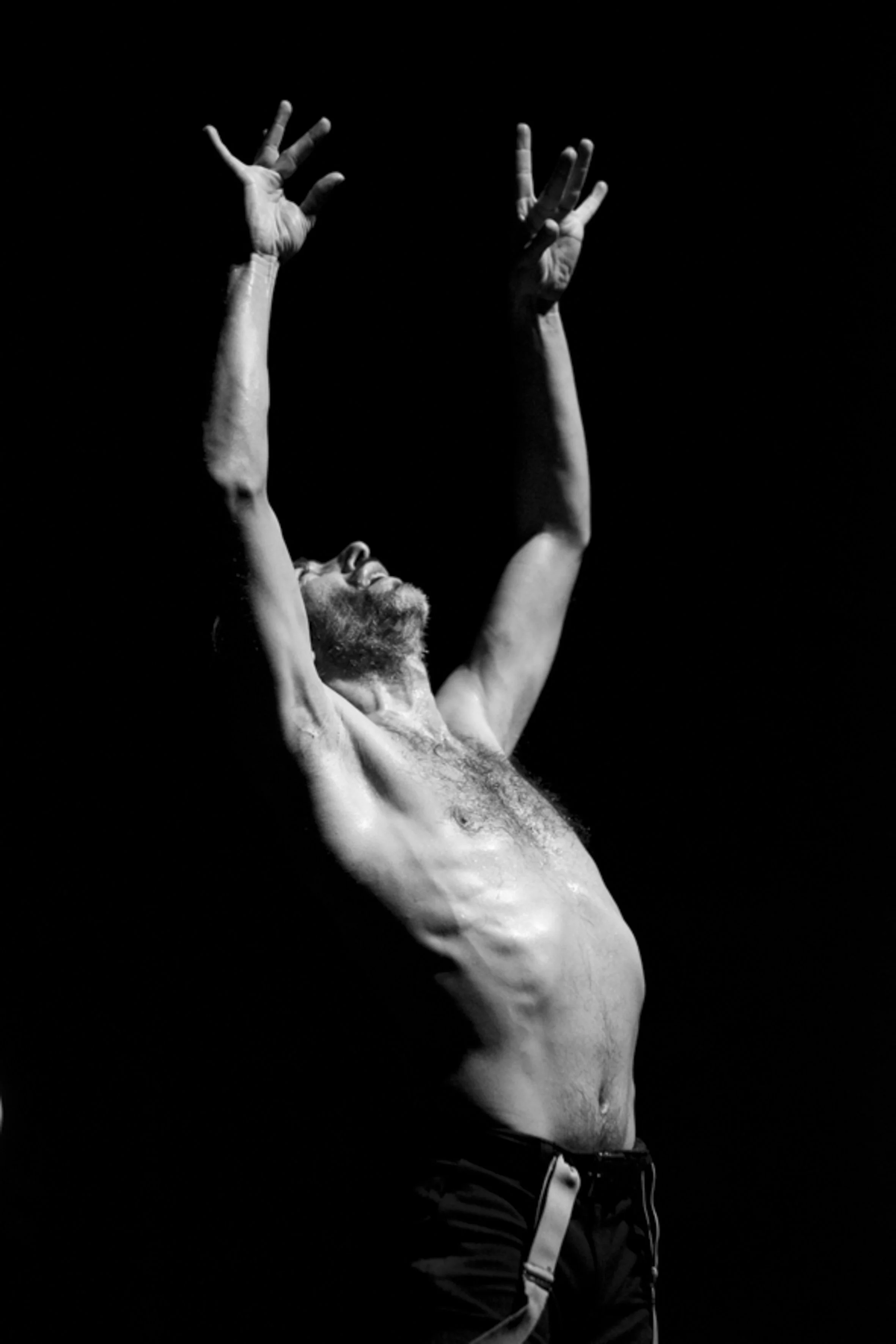Homme nu avec barbe, bras levés, en position expressive contre un fond noir.