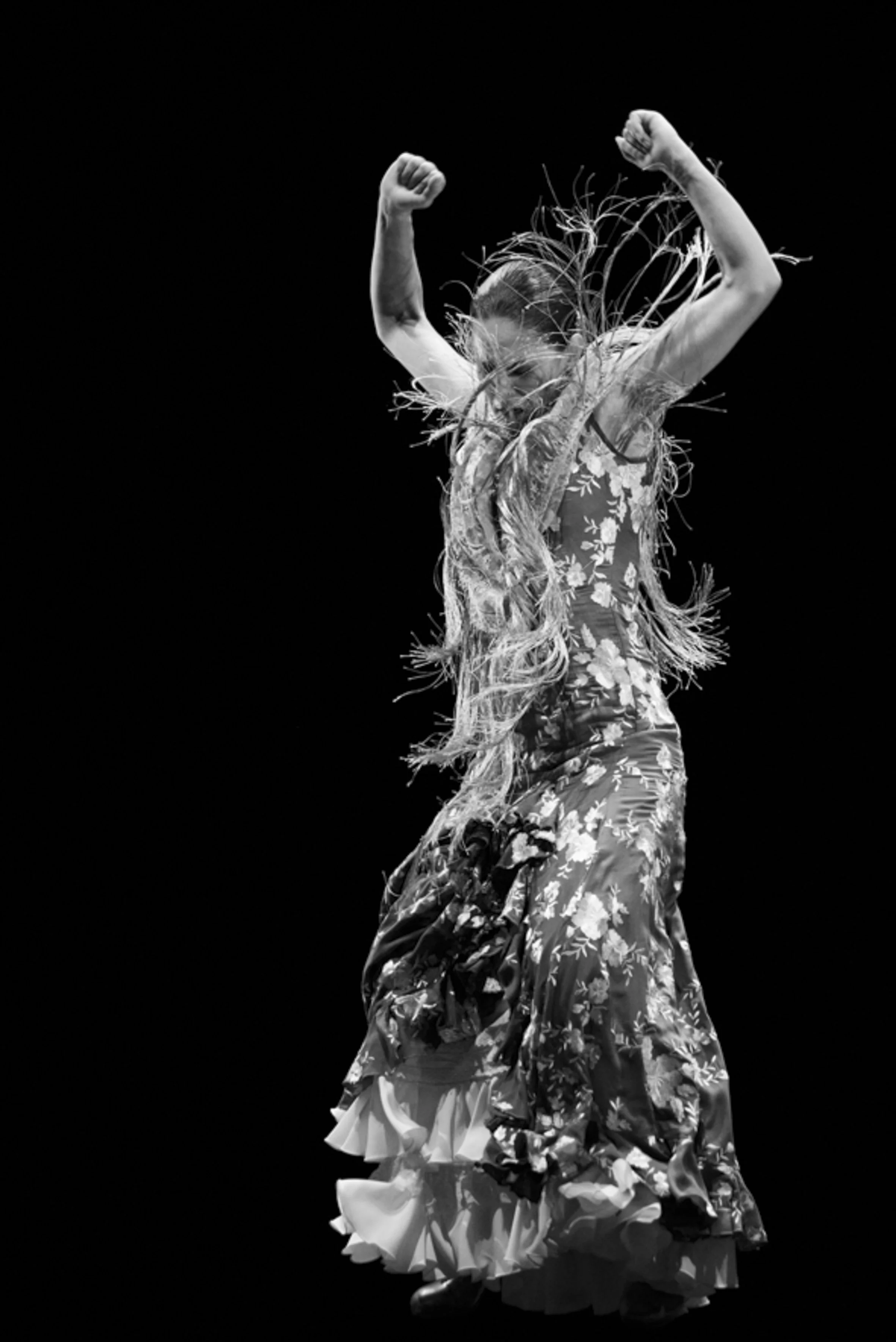 Une femme danse portant une robe longue à motifs floraux, avec les bras levés et un fond noir.