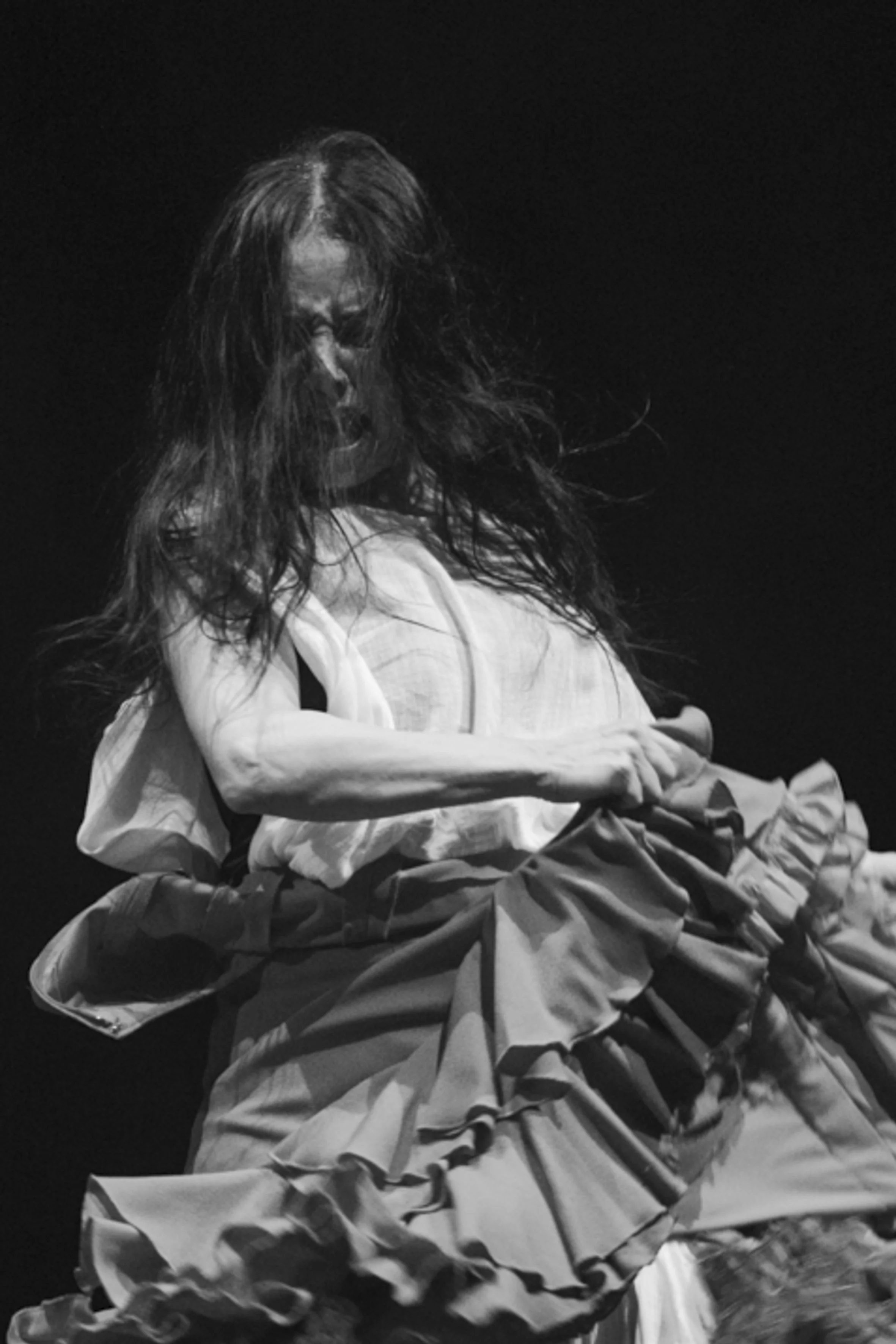 Une femme en mouvement, portant une robe volumineuse, avec cheveux longs et bouclés, exprimant une émotion intense, en noir et blanc.