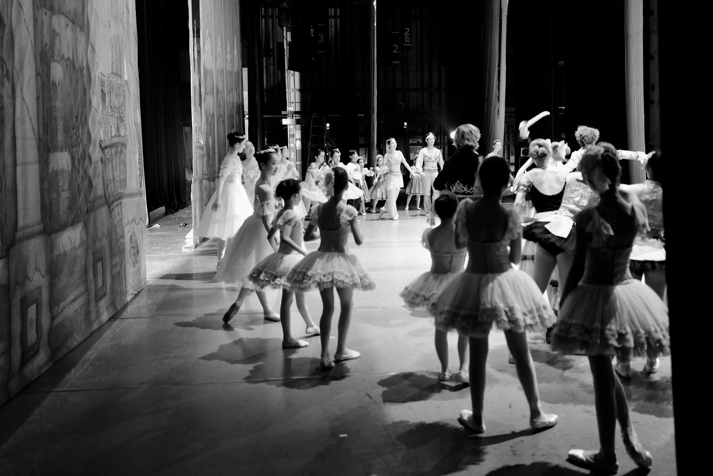 Ballet dancers practicing on stage, many wearing tutus and ballet costumes, with an instructor guiding them in a dance rehearsal.