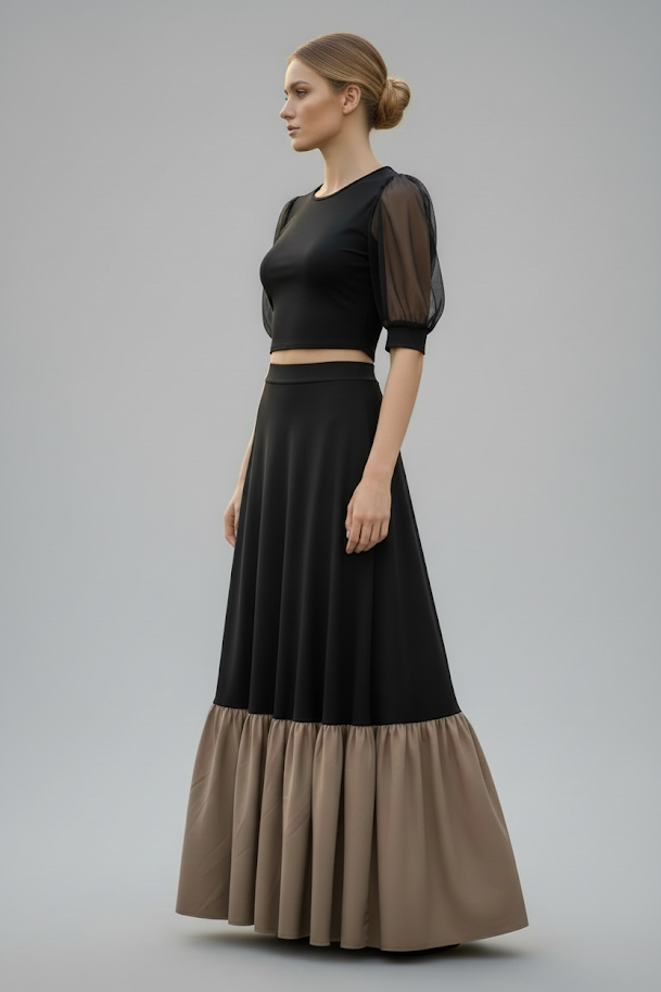A woman with blonde hair tied in a low bun wearing a black crop top with sheer puff sleeves and a long black and beige ruffled skirt standing sideways against a plain gray background.