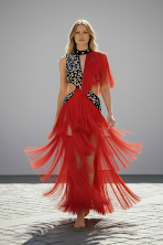A model walking down the runway wearing a vibrant red fringe dress with a black and white patterned top and long blonde hair.