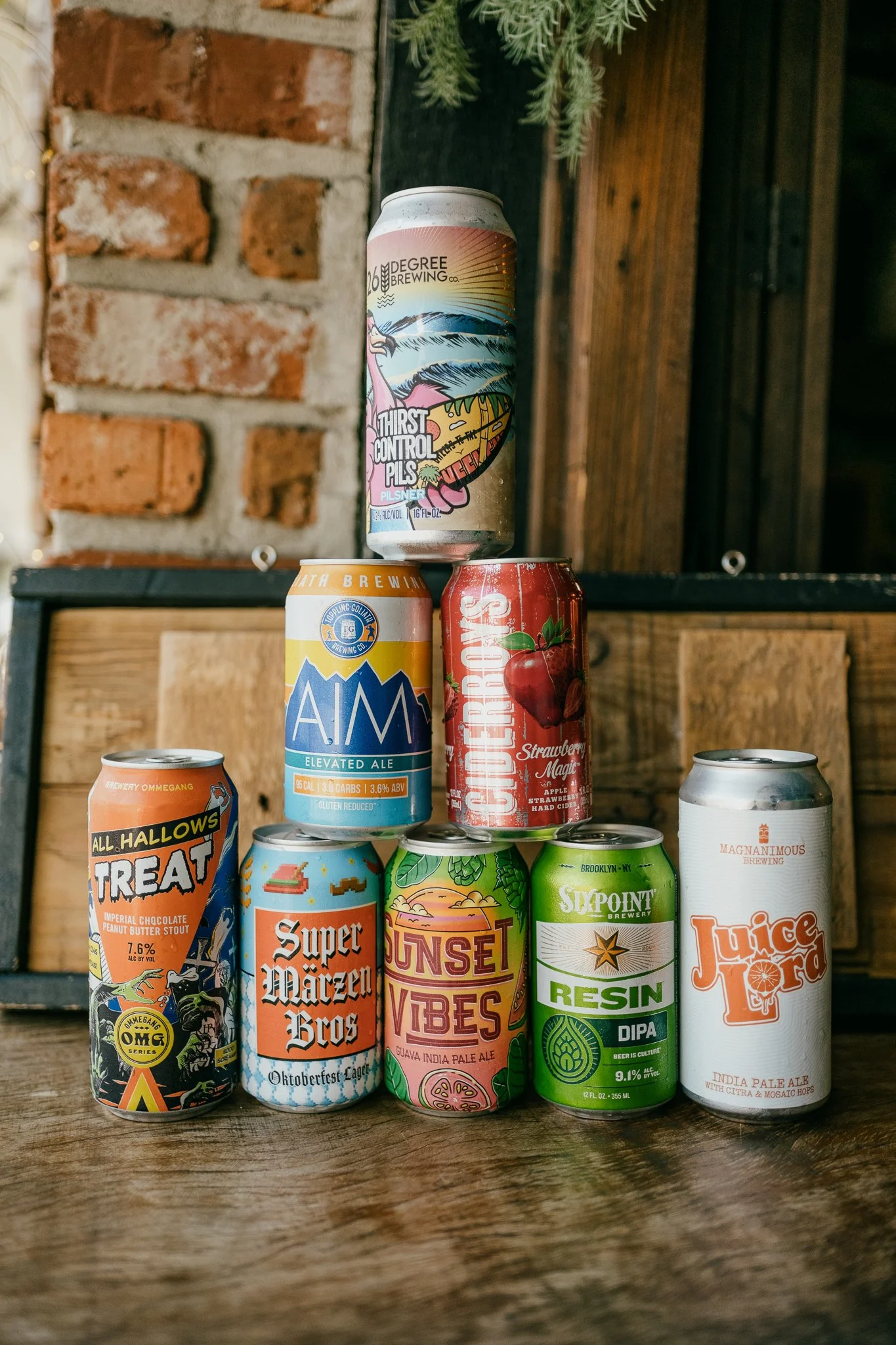 Six cans of different craft beers arranged on a rustic wooden table, with a brick wall and wooden paneling in the background.