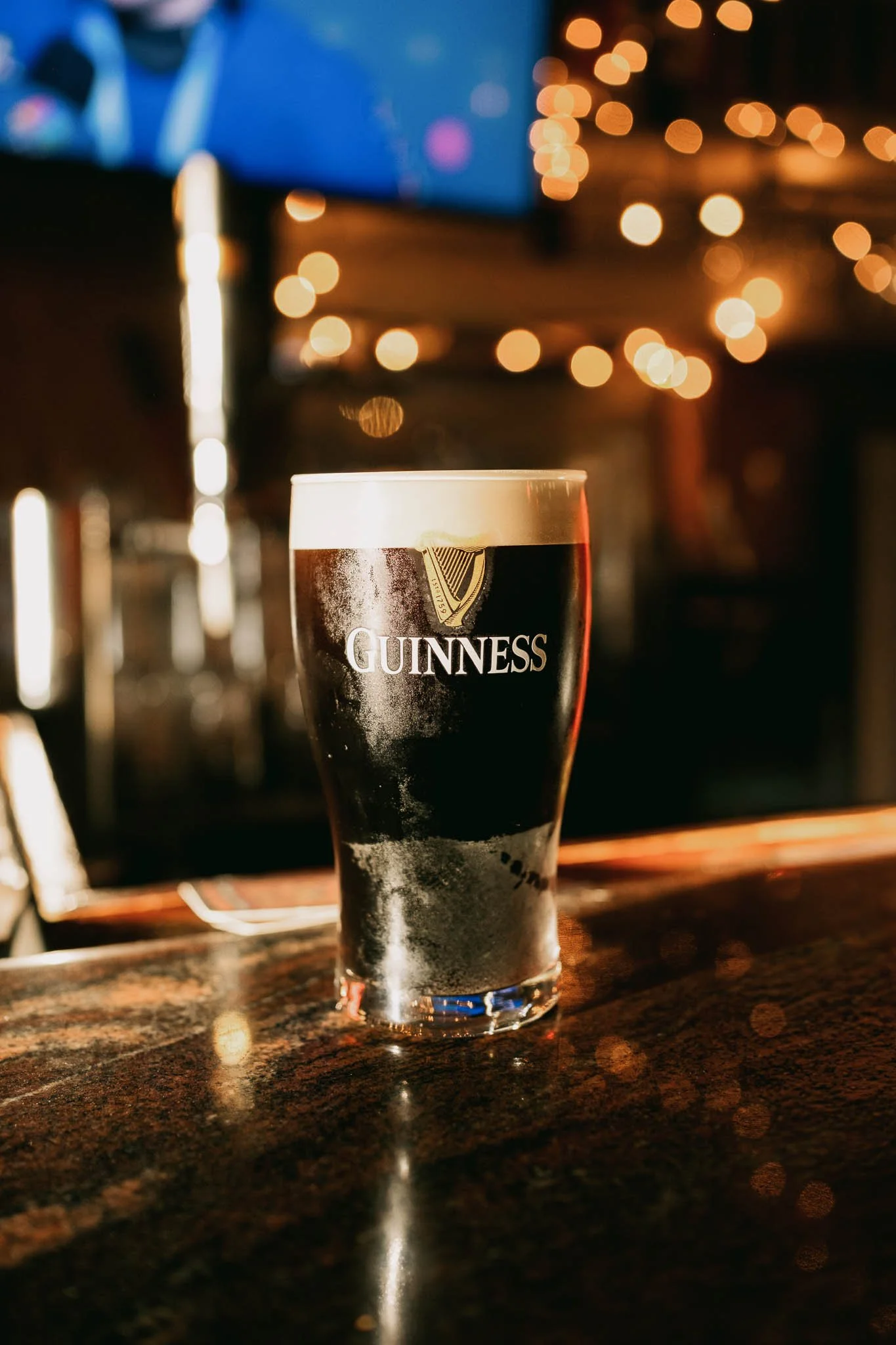 A pint of Guinness beer on a bar counter at night with blurred, warm-colored lights in the background.