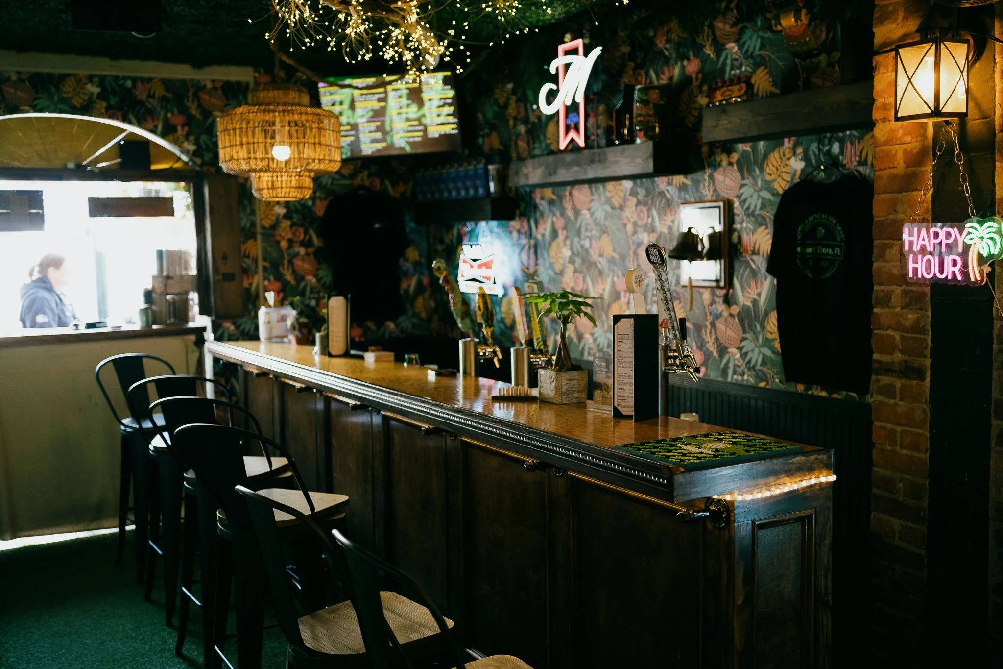 Interior of a bar with tropical-patterned wallpaper, a long wooden counter, and black chairs. Decorative lighting, neon signs, and a woman at the window are visible.
