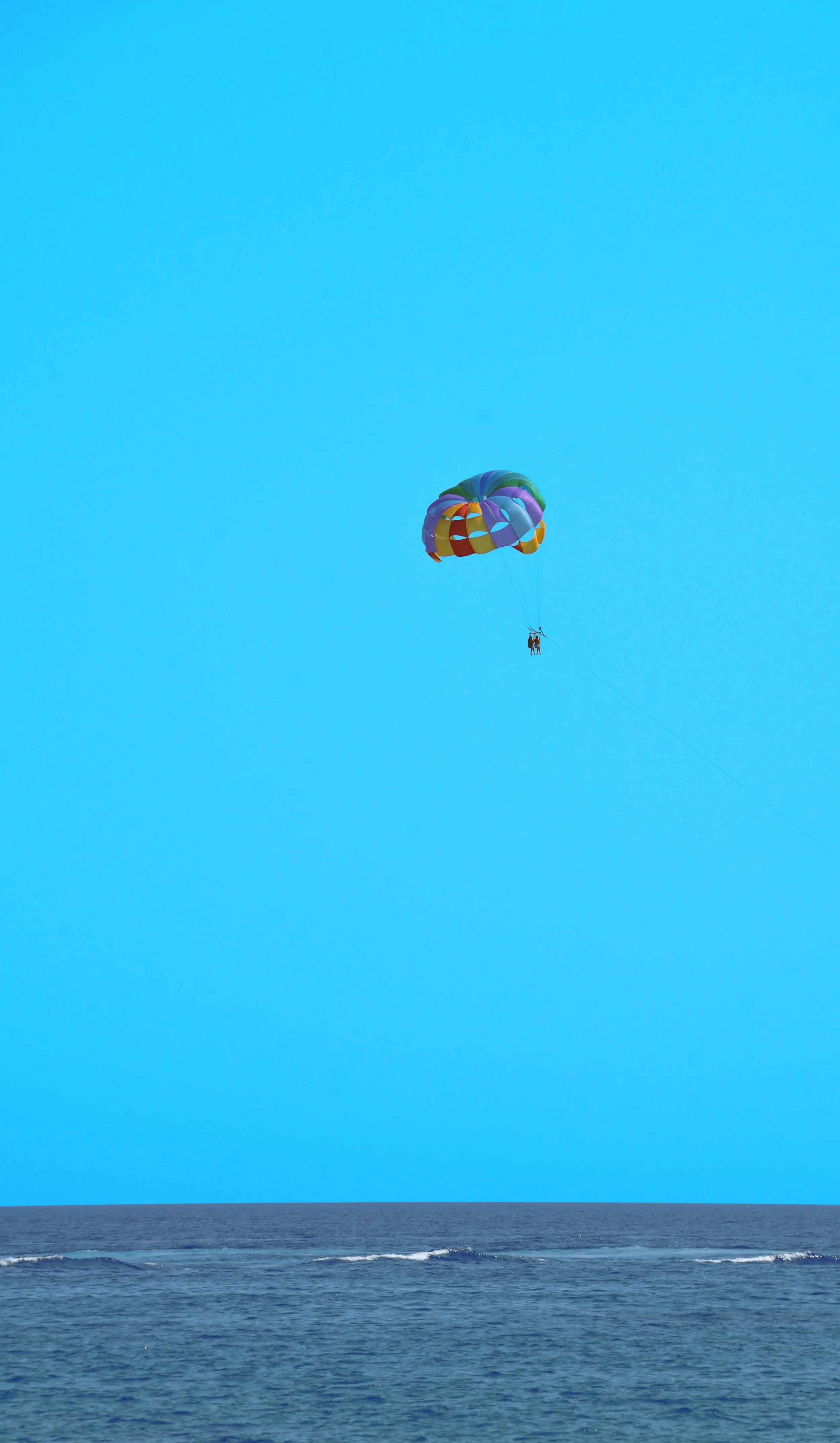 Minimalist vertical photograph dominated by flat cyan sky. A multicolored parasail with two suspended figures centered in the upper third of the frame. A thin dark strip of open ocean with faint white waves occupies the lower fifth.