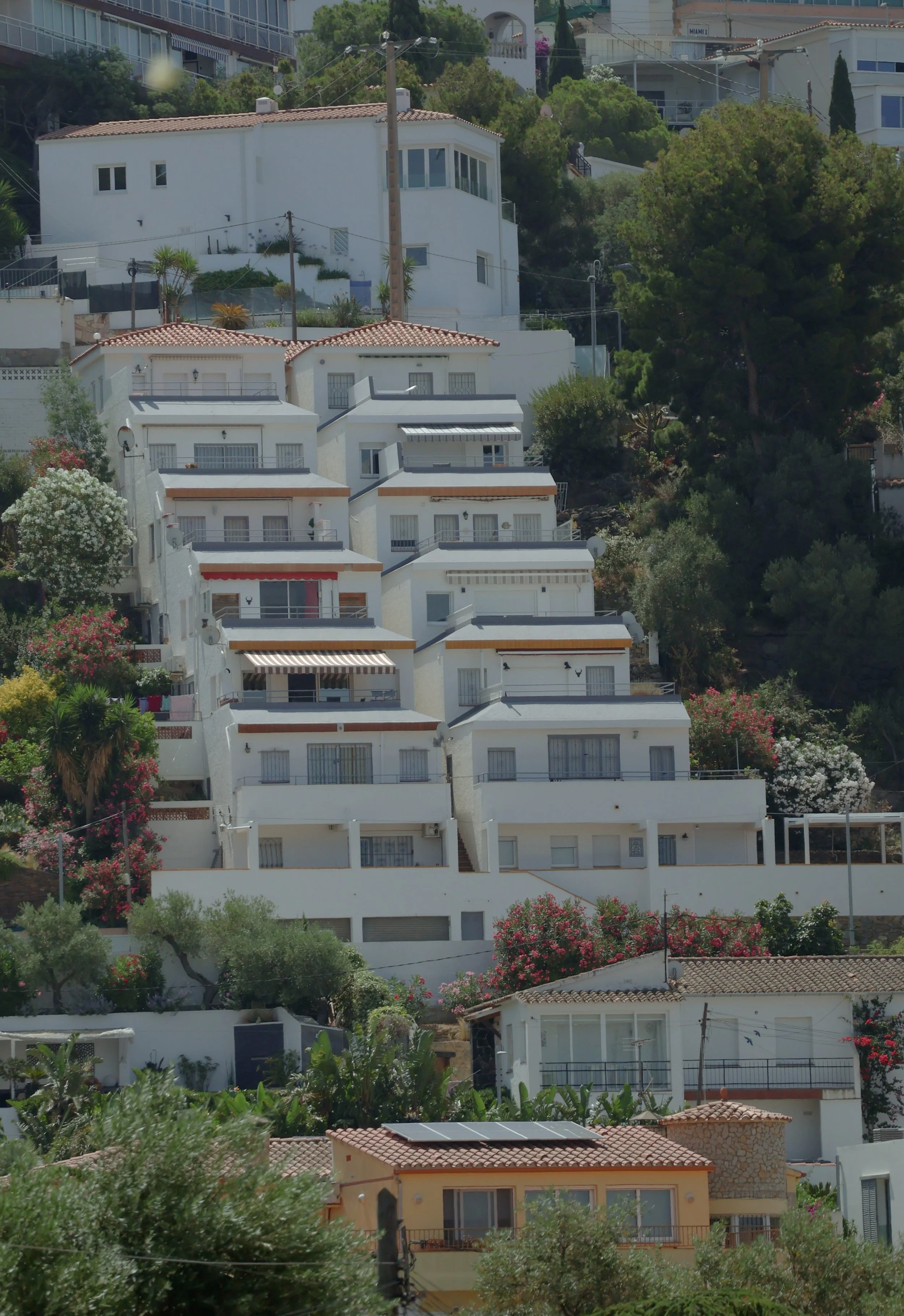 Telephoto compression of a white multi-storey apartment complex built in stepped terraces down a Mediterranean hillside. Repeated balconies and windows in near-identical rhythm, punctuated by a red awning and a striped canopy.
