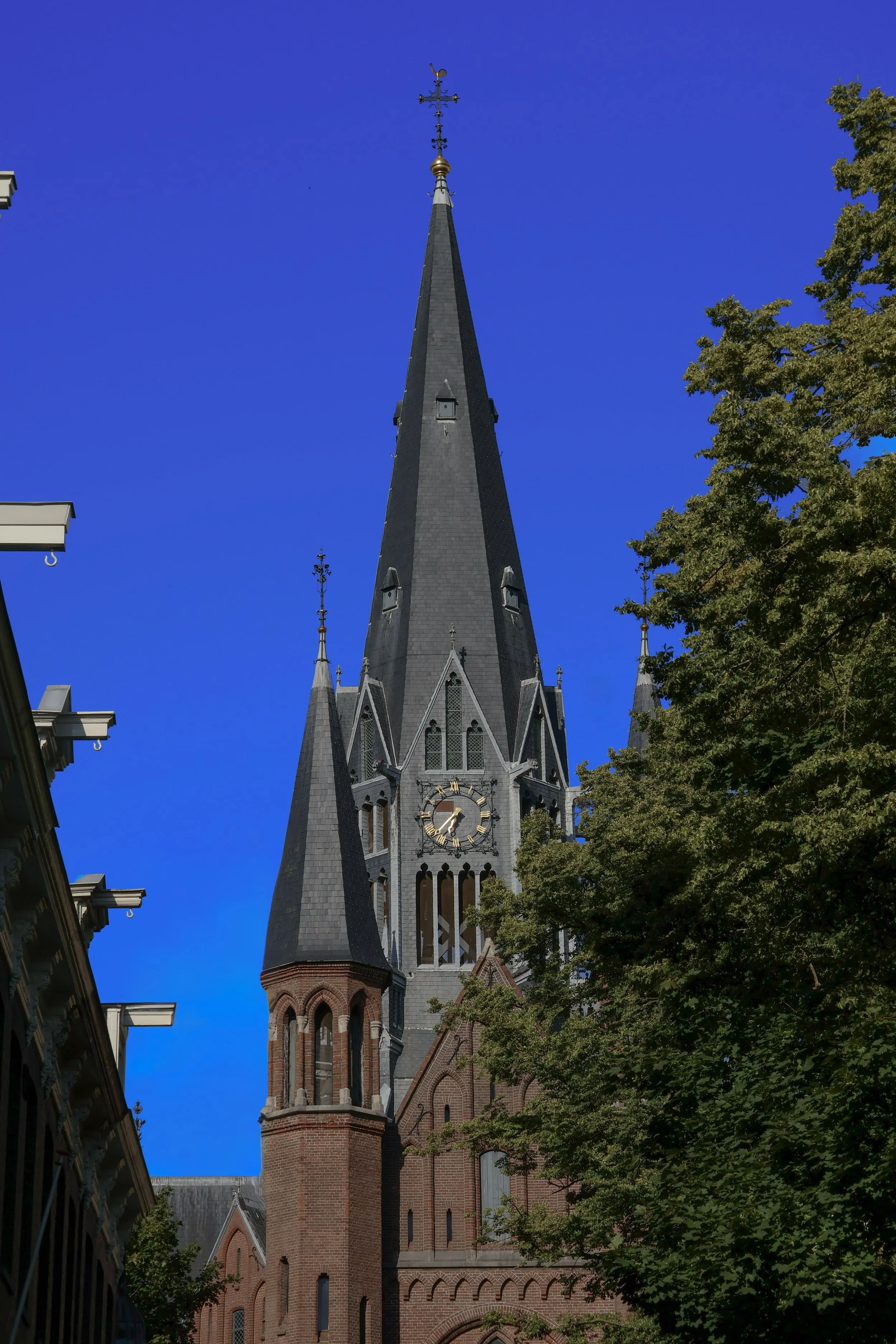 Low-angle view of a Neo-Gothic red brick church tower and slate spire, featuring a gold Roman numeral clock face, ornate Gothic lancet windows, decorative pinnacles and iron cross finial