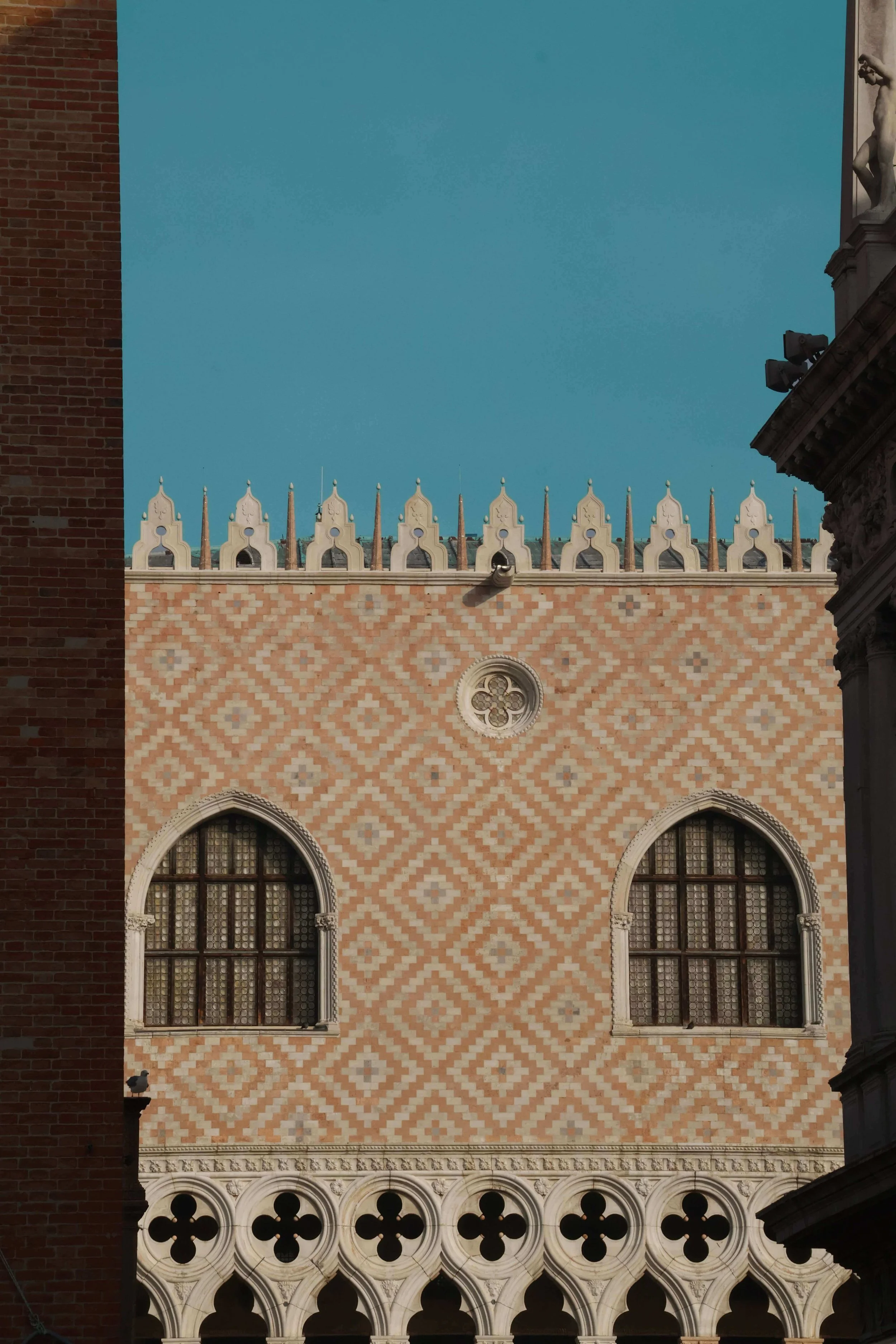 Frontal architectural detail of the Doge's Palace upper facade in Venice. Pink and white diamond-pattern marble wall with two Gothic arched windows and a central rose medallion.