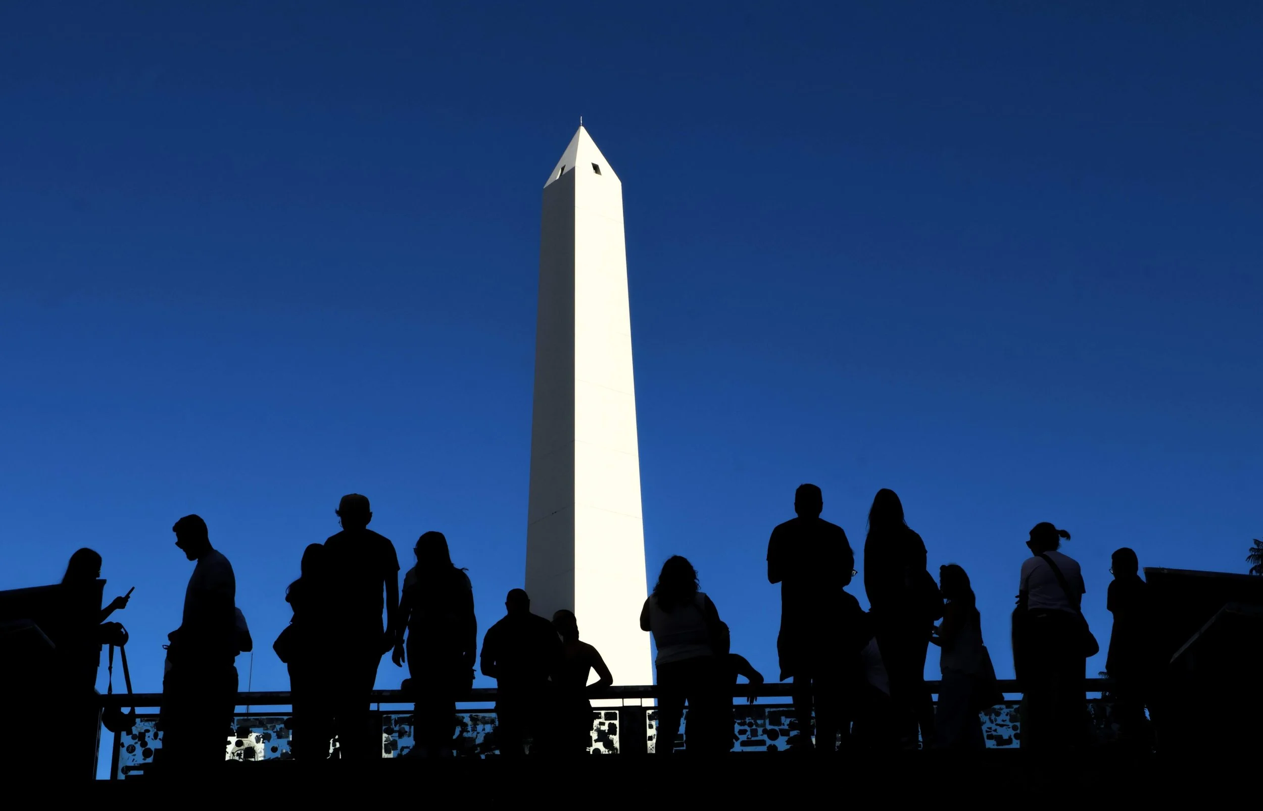 High contrast photograph of the Obelisco de Buenos Aires — white stone monument against a deep cobalt blue midday sky. A row of black silhouetted figures leaning on a railing in the foreground, none facing the camera.