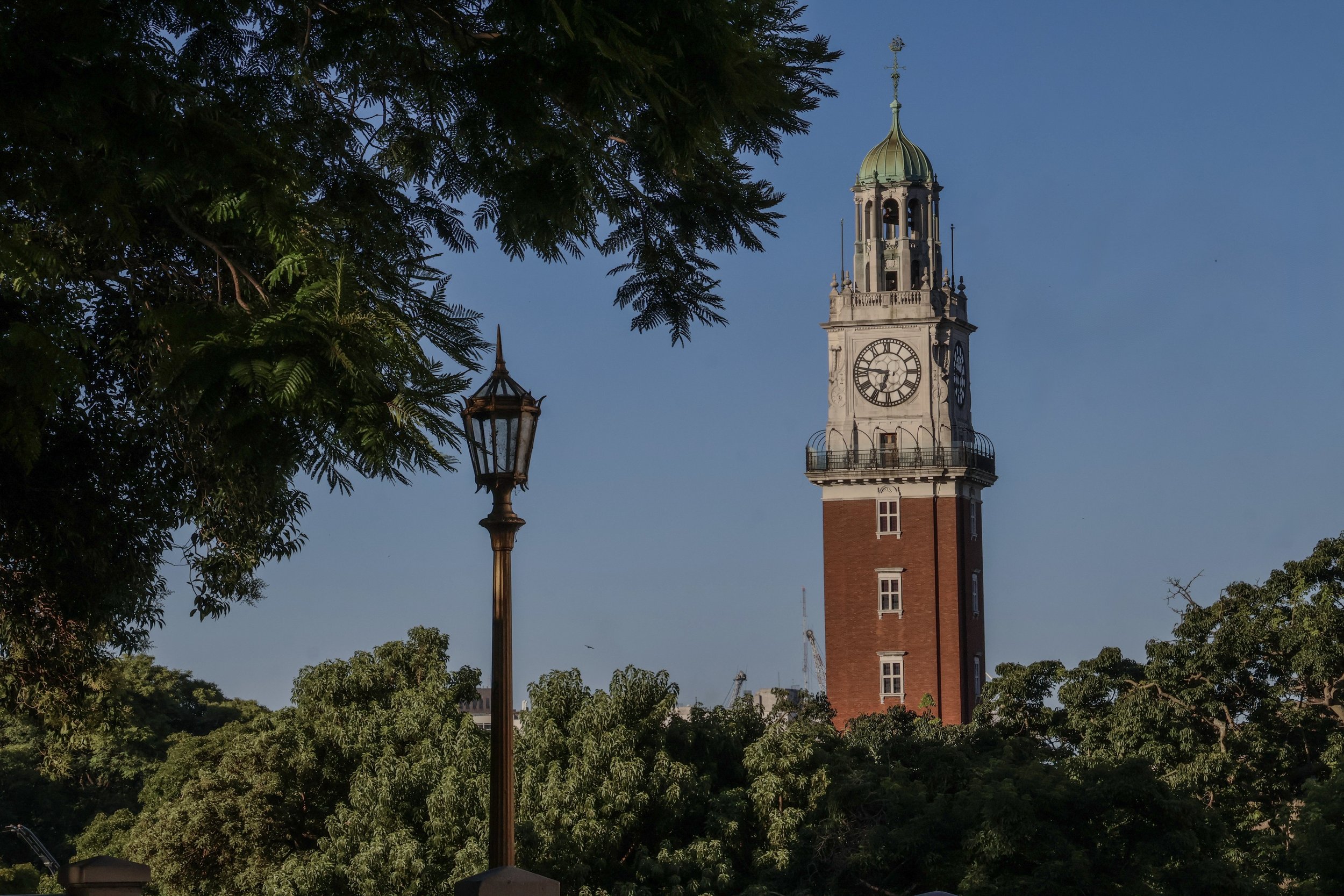 Fine Art photography of the Monumental Tower in Buenos Aires at 18:46. Red brick and white stone clock tower framed by green tree foliage and a vintage street lamp under a clear blue sky