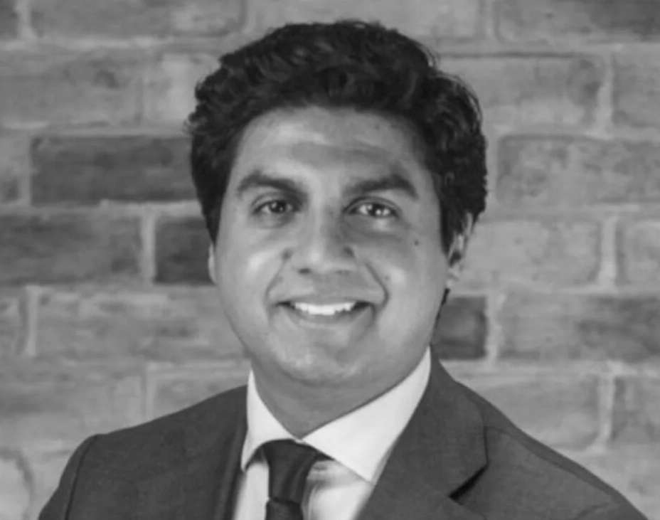 Black and white photo of a man in a suit and tie, smiling, with a brick wall background.