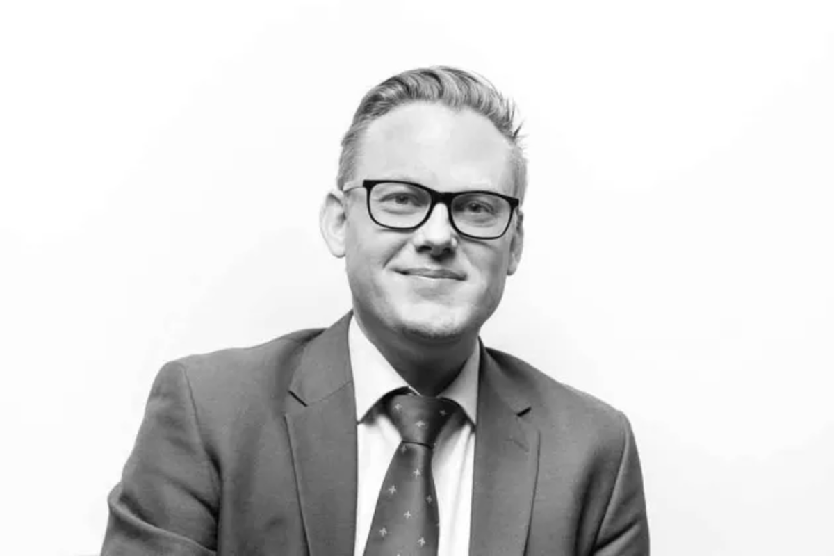 Black and white portrait of a man in a suit and tie, wearing glasses, smiling, against a plain light background.