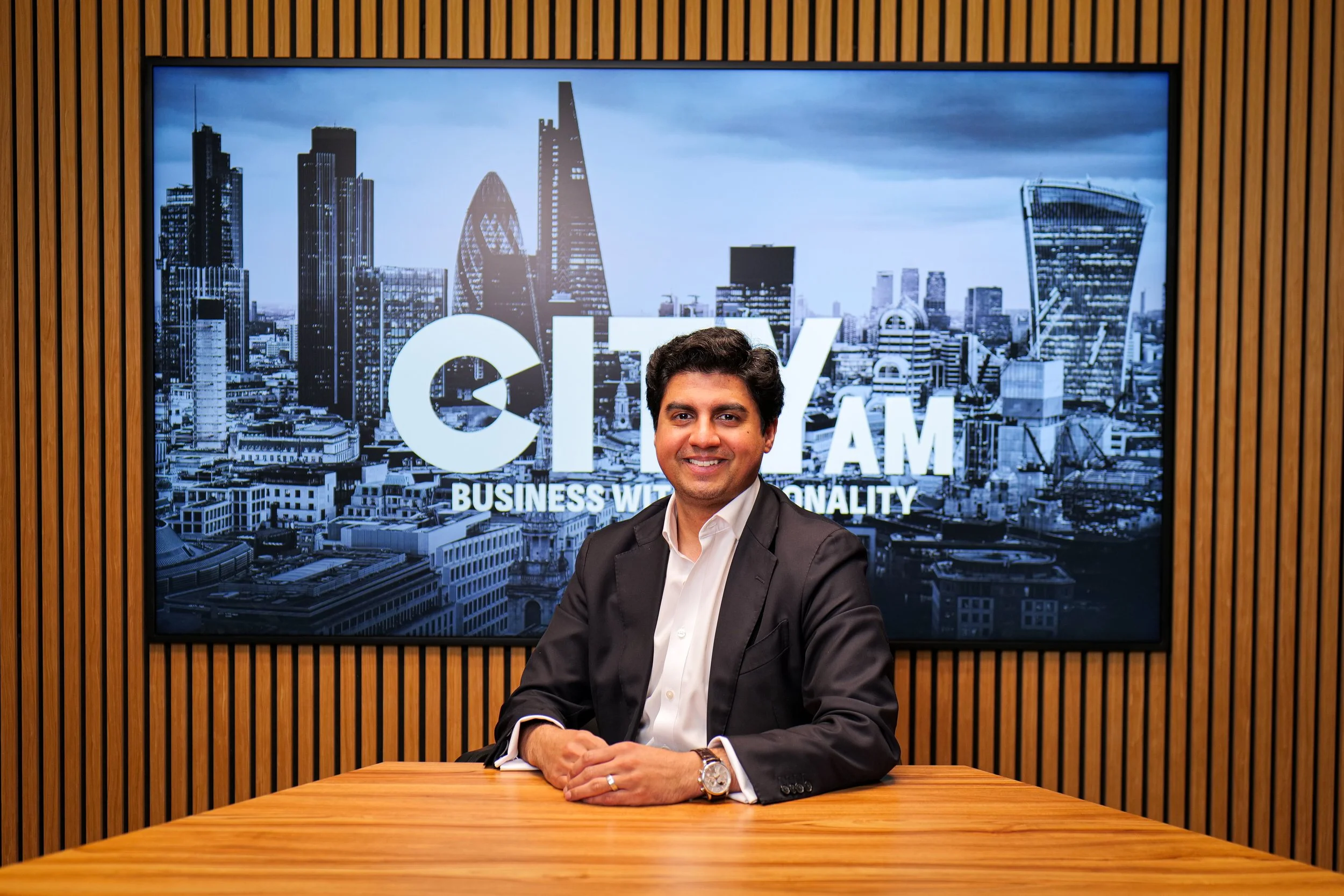 Black and white photo of a man in a suit and tie, smiling, with a brick wall background.